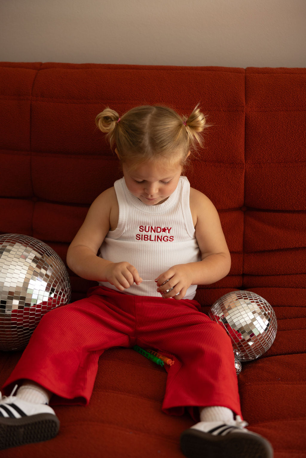 A young child with blonde hair in pigtails sits on a red sofa, wearing a white PRE-ORDER Minnie Singlet by SUNDAY SIBLINGS and Supersonic Cord Pants made of ribbed cotton fabric. Nearby are two disco balls. The child is focused on a small object in their hands.