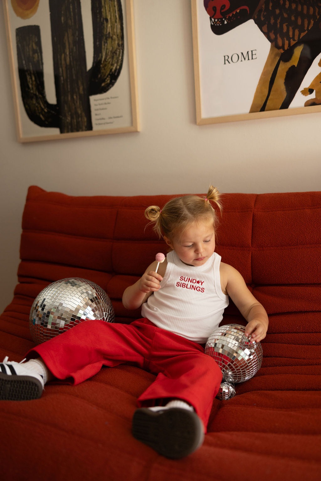 A child with blonde hair in pigtails sits on a red couch, wearing a white PRE-ORDER Minnie Singlet by SUNDAY SIBLINGS and red Supersonic Cord Pants. They hold a lollipop and are surrounded by disco balls, while two framed artworks hang on the wall behind them.