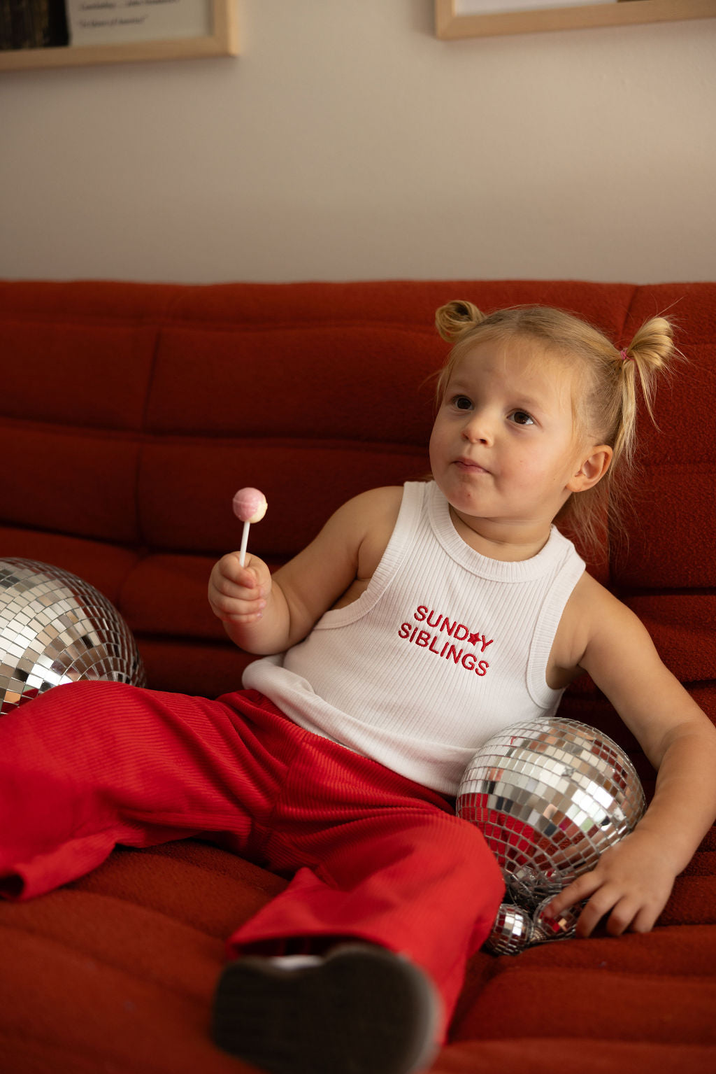 A child with two small ponytails sits on a red sofa holding a pink lollipop. They wear the PRE-ORDER Minnie Singlet from SUNDAY SIBLINGS paired with vibrant Supersonic Cord Pants. Two disco balls glisten on either side of the scene, completing the cheerful picture.