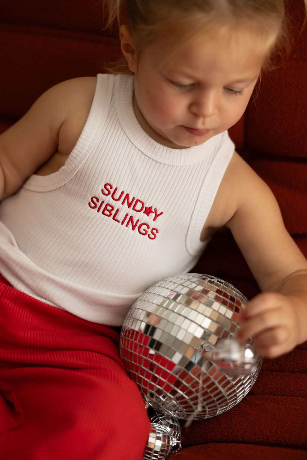 A child wearing the PRE-ORDER Minnie Singlet by SUNDAY SIBLINGS, paired with red Supersonic Cord Pants, sits on a matching red chair. They hold a small disco ball, gazing at it attentively. The outfit is completed with an embroidered logo, adding subtle charm.