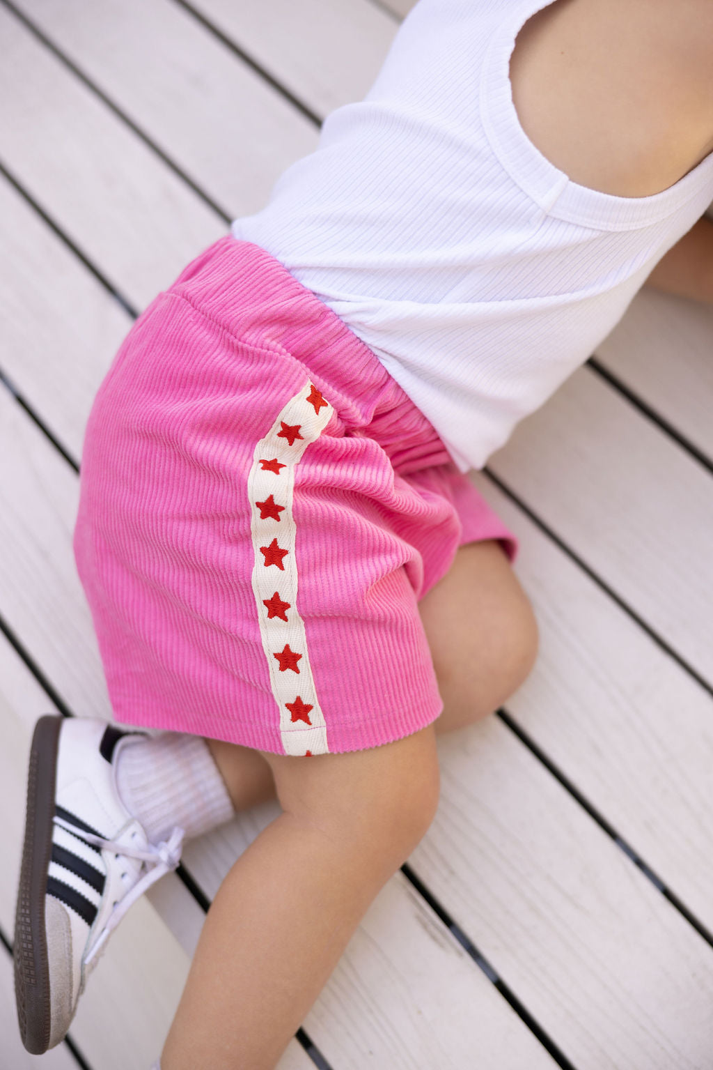 A child wearing a white tank top and SUNDAY SIBLINGS' Playtime Cord Shorts in pink, adorned with star embroidery, crawls on a white wooden surface. The child also sports white sneakers with black stripes, effortlessly merging comfort and style during their playful adventure.