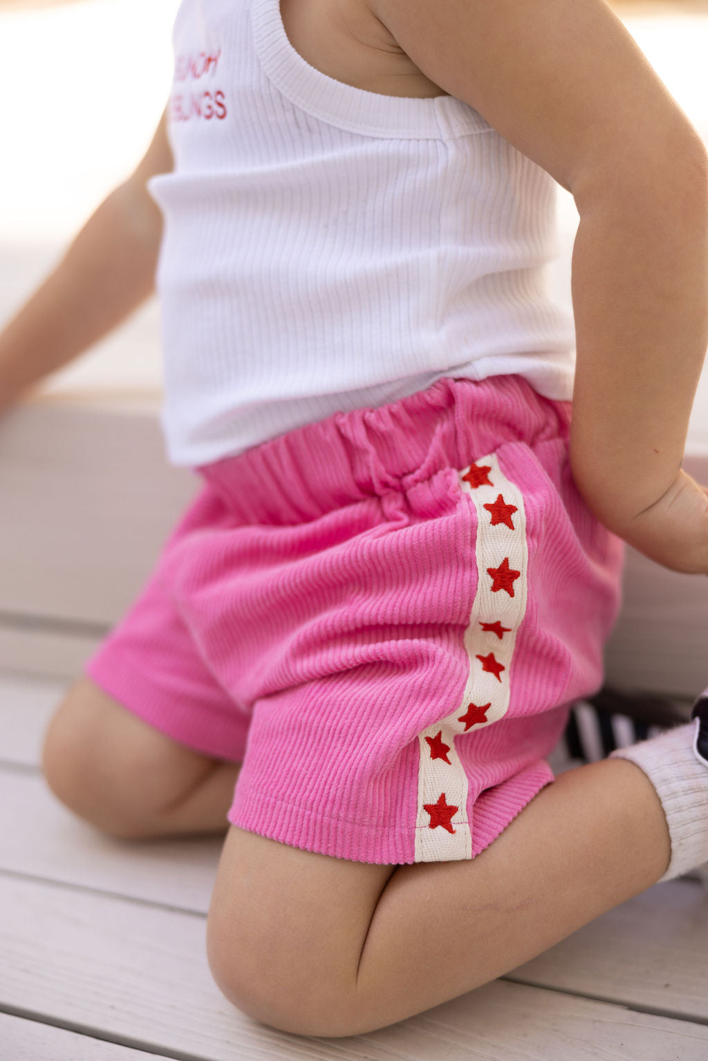 A child wearing a white sleeveless top and SUNDAY SIBLINGS Playtime Cord Shorts Pink with star embroidery sits with one knee on the ground. The shorts, featuring a white ribbon with red stars, complement the child's look. Their hand rests on the floor as they sport a black shoe and white sock.