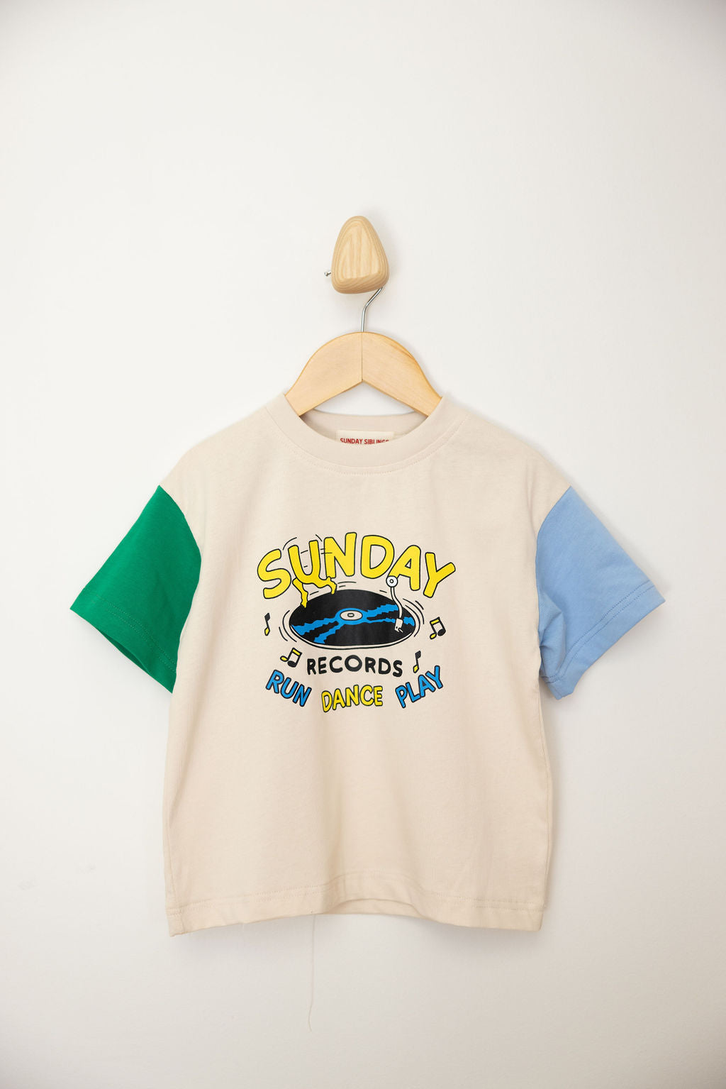 A children's T-shirt from SUNDAY SIBLINGS, the Sunday Records Tee Blue/Green, hangs on a wooden wall hook. It features multicolored sleeves—one in green and the other in blue. The front showcases a phonograph alongside the text "Sunday Records Run Dance Play," epitomizing the signature Marcus Dixon style with its oversized fit.