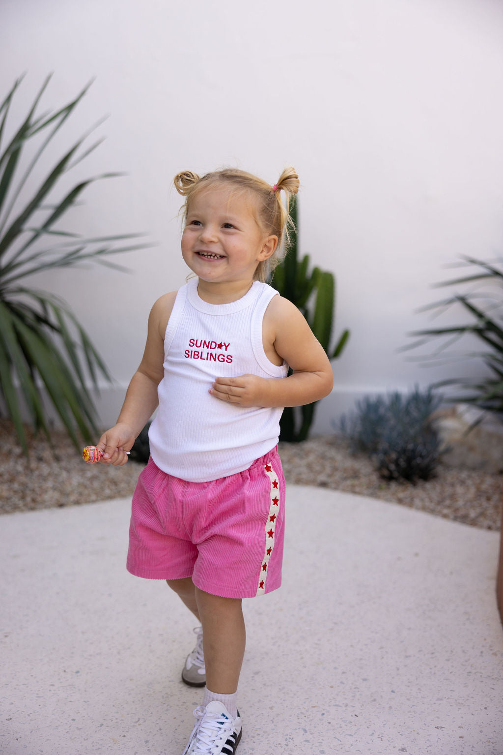 A cheerful toddler with blonde pigtails is wearing the PRE-ORDER Minnie Singlet by SUNDAY SIBLINGS, a white sleeveless top made of ribbed cotton fabric. She pairs it with pink shorts adorned with star details while holding a lollipop and standing outdoors amidst greenery.