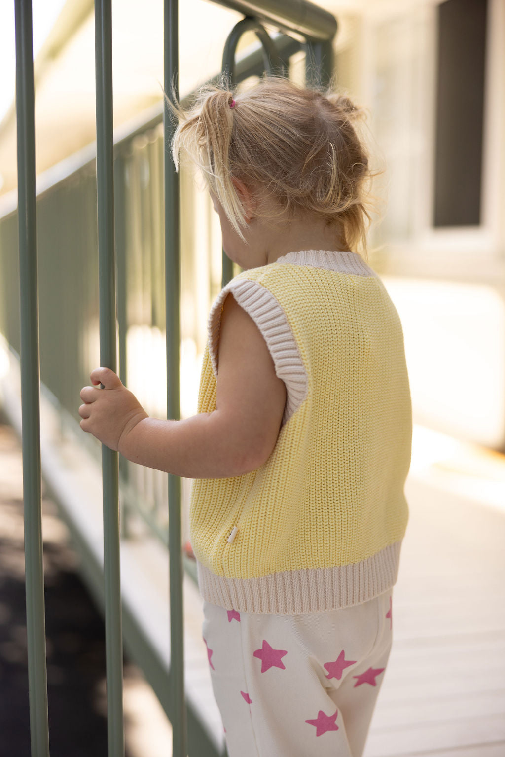 A young child with blonde pigtails stands on a porch, holding onto a green railing. They are wearing the SUNDAY SIBLINGS Nonno Vest in butter yellow, made from 100% cotton, along with white pants adorned with pink stars, all while facing away from the camera.