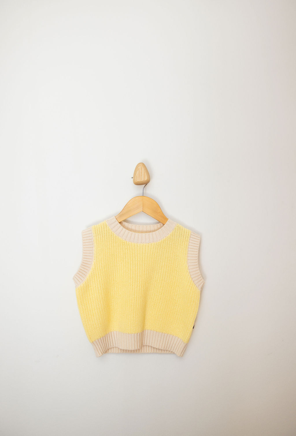 The Nonno Vest Butter Yellow by SUNDAY SIBLINGS, featuring a sleeveless design crafted from 100% cotton with beige trim, hangs elegantly on a wooden hanger against a plain white wall.