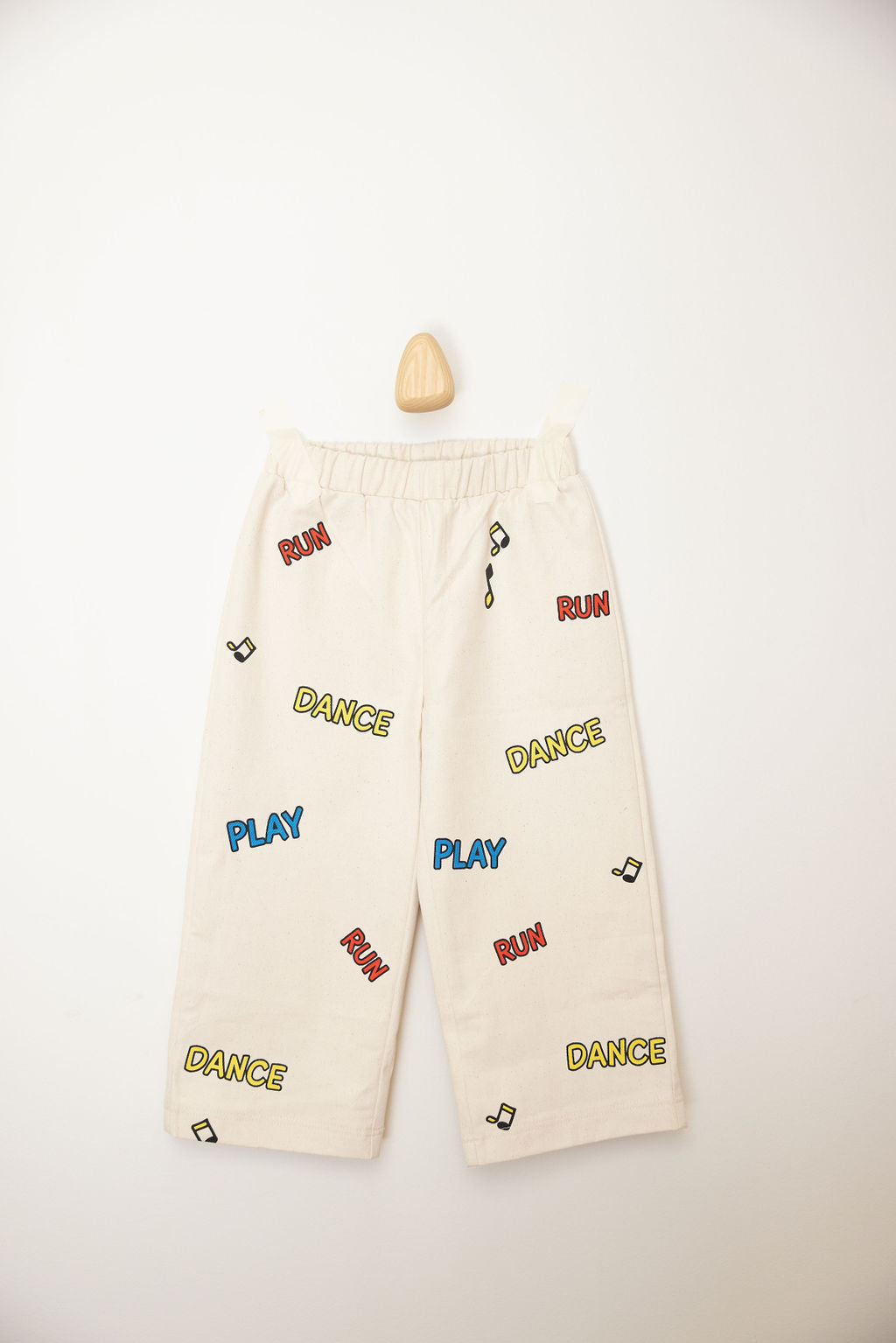 The Sunday Records Pants by SUNDAY SIBLINGS are white children's pants made from soft twill cotton. They feature colorful text prints in red, blue, and yellow that say "RUN," "PLAY," and "DANCE." The design is complemented by small black music notes. These pants include an elastic waistband for comfort and come with patch pockets, displayed flat against a light background.