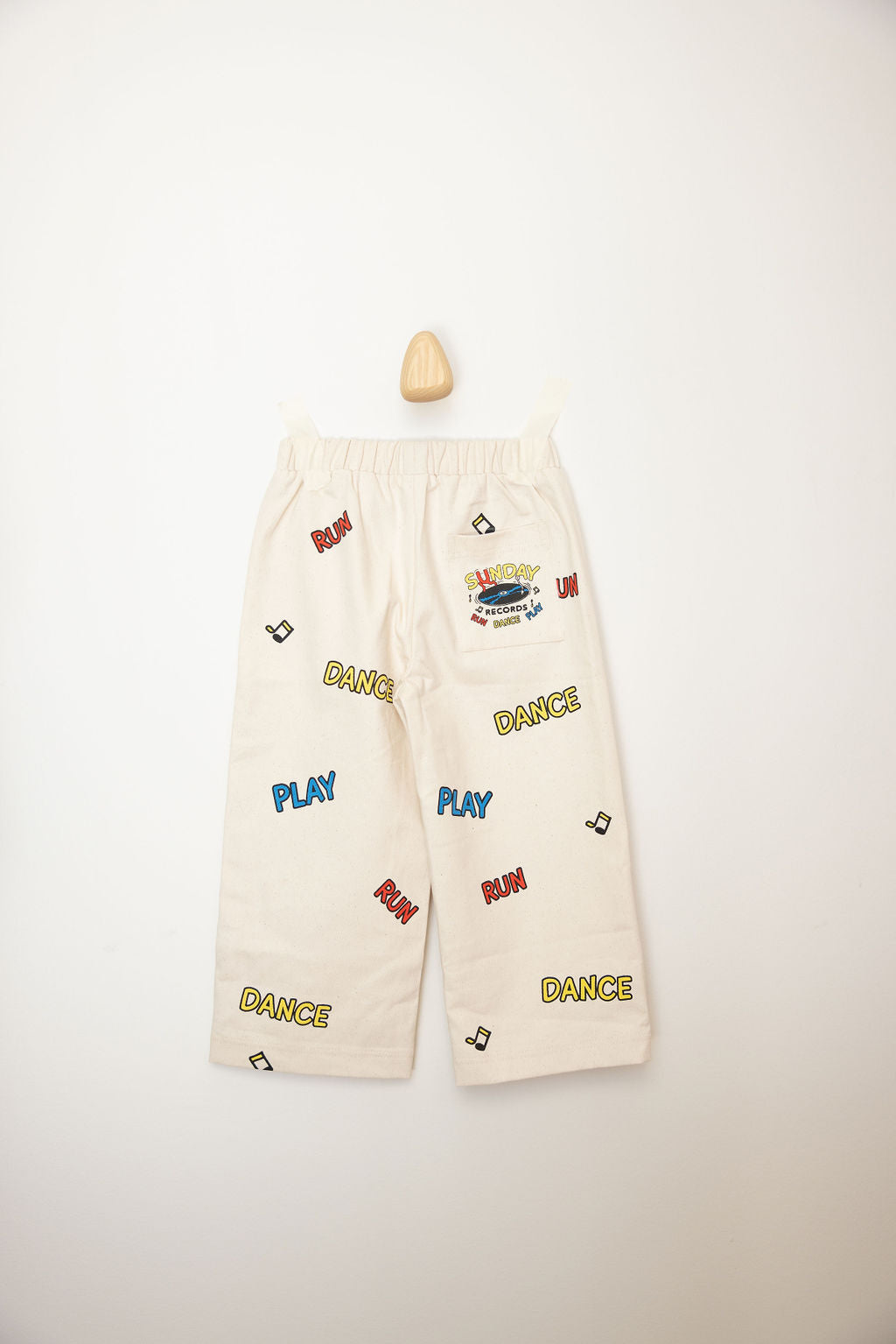 The Sunday Records Pants by SUNDAY SIBLINGS are beige children's pants made from twill cotton, adorned with vibrant printed words such as "PLAY," "DANCE," and "RUN." These pants feature an elastic waistband for comfort and are presented flat against a neutral background, complete with a small wooden hanger above.