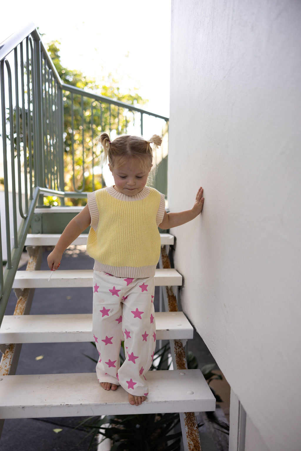 A child with pigtails, in a yellow sleeveless top and SUNDAY SIBLINGS Ninch Pants Pink Stars (Size 5-6) with an elastic waistband, walks down a metal outdoor staircase on a sunny day, holding the railing amid visible greenery.