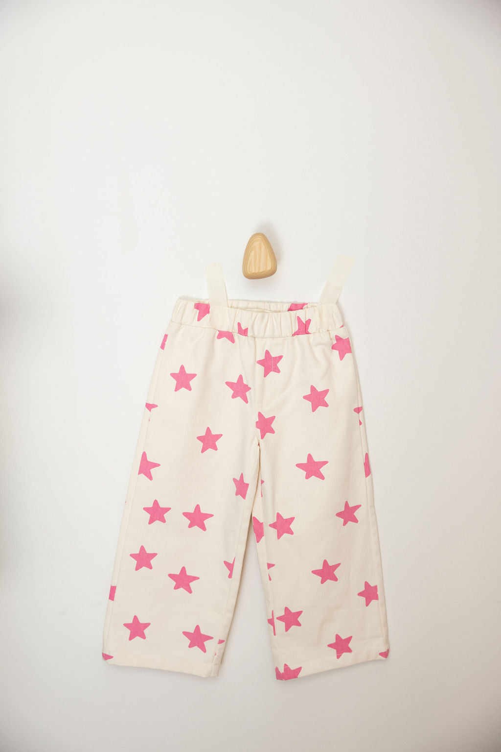 The PRE-ORDER Ninch Pants Pink Stars from SUNDAY SIBLINGS, crafted from soft twill cotton and featuring pink star patterns, are displayed on a wall against a plain background.