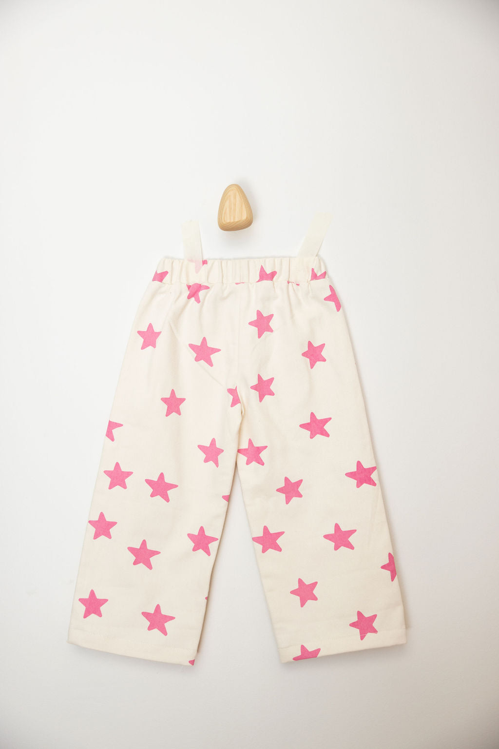 The PRE-ORDER Ninch Pants Pink Stars by SUNDAY SIBLINGS, crafted from soft twill cotton and decorated with pink star patterns, hang on a wall against a light background. A wooden hook is visible at the top, showcasing their elastic waistband. Ideal for style-savvy tots, these pants are now available for pre-order.