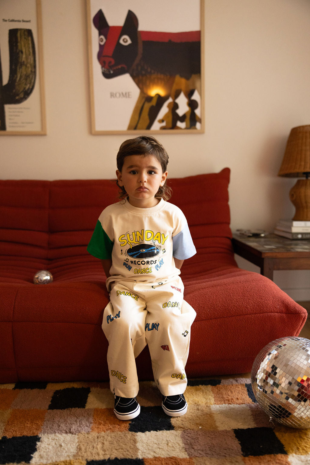 A young child with long hair sits on a red couch in a living room, wearing an oversized SUNDAY SIBLINGS Sunday Records Tee Blue/Green with colorful graphics and black shoes. Near the disco ball on the floor, the rug boasts geometric patterns, creating a vibrant scene reminiscent of something Marcus Dixon might capture.