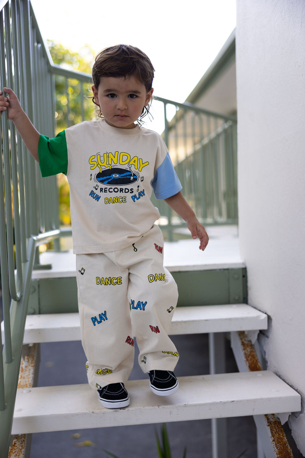 A young child stands on the stairs, holding a railing in their oversized Sunday Siblings Sunday Records Tee Blue/Green. Wearing colorful patterned pants emblazoned with "Dance" and "Play," they exude joy against a backdrop of greenery and part of a building in this charming scene captured by Marcus Dixon.