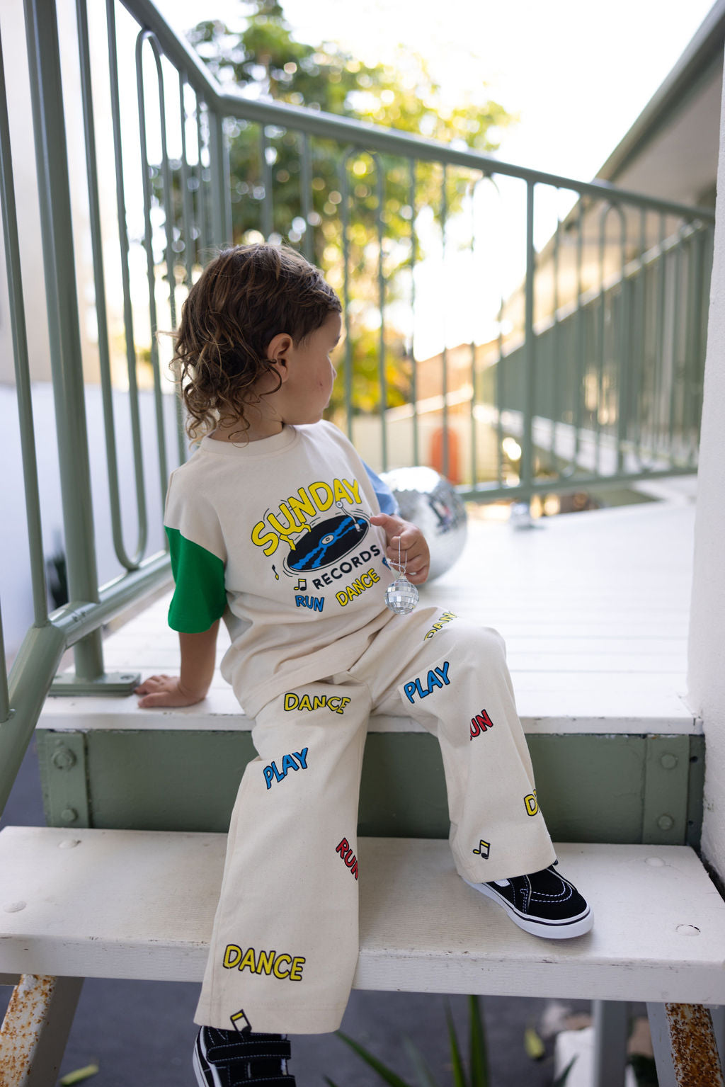 A young child with curly hair sits on outdoor steps, their outfit capturing the essence of playful fashion. They're wearing the Sunday Records Pants from SUNDAY SIBLINGS, featuring a "Sunday" print over a vinyl record graphic on the shirt and complemented by matching pants adorned with "Dance" and "Play." The twill cotton pants come with patch pockets and an elastic waistband, while a silver disco ball glimmers nearby.