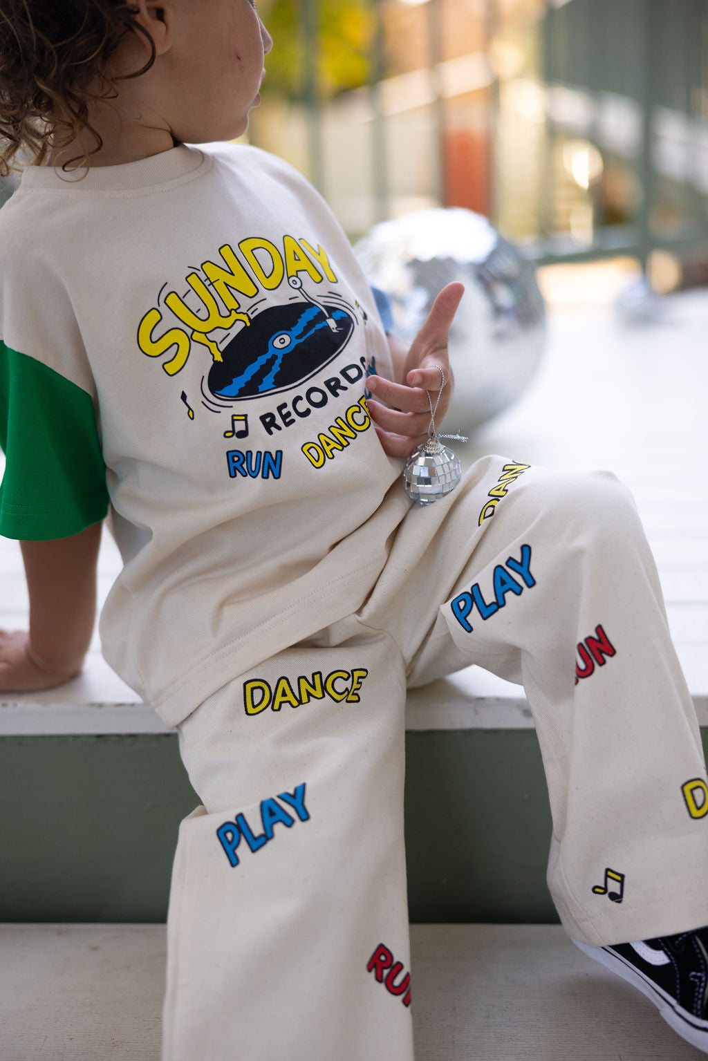 A child sits on a step, wearing a colorful ensemble of Sunday Records Pants by SUNDAY SIBLINGS. The twill cotton pants feature an elastic waistband and patch pockets, adorned with words like "Sunday," "Record," "Run," "Dance," and "Play." They hold a small, shiny object in one hand, with a large metallic sphere visible behind them.