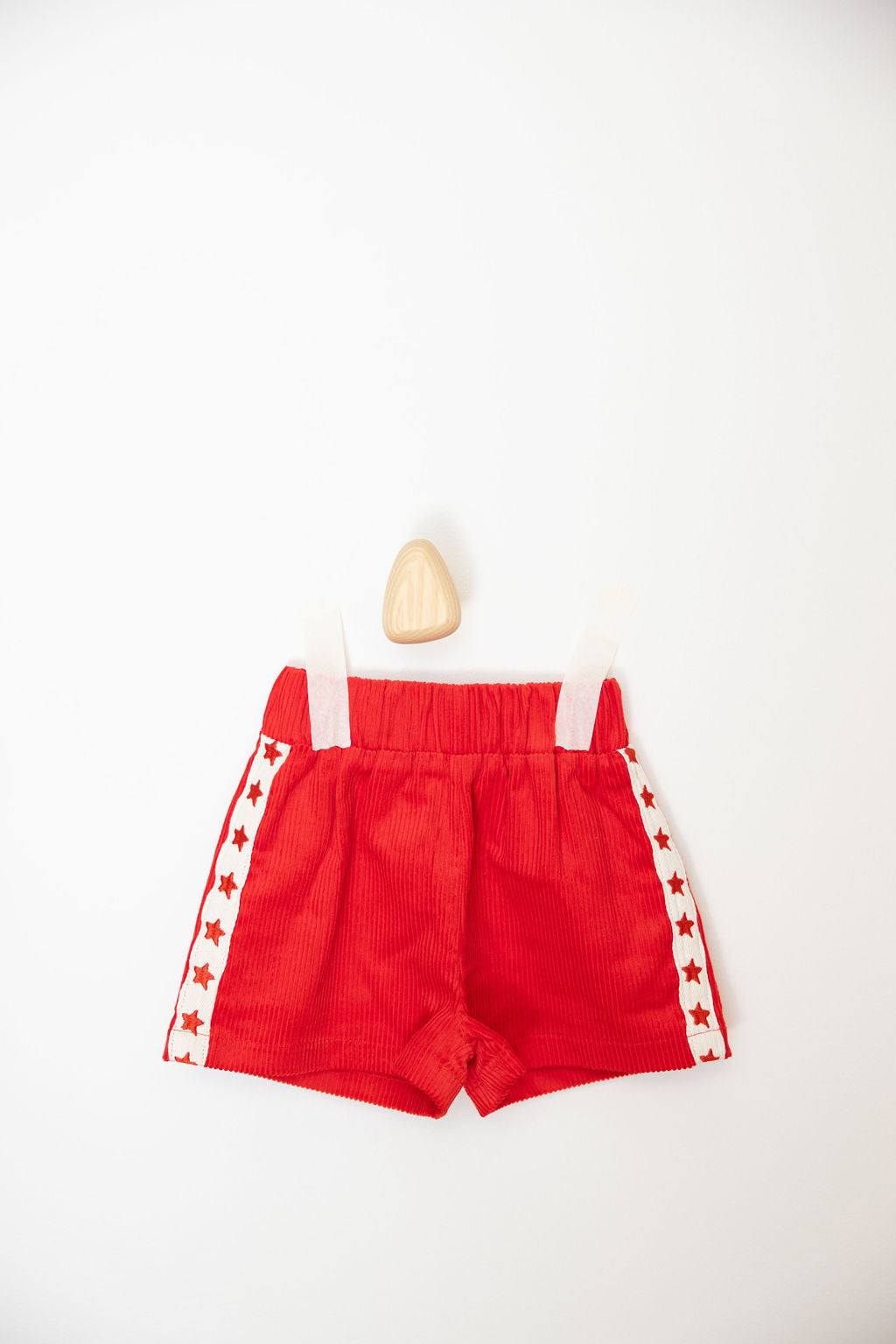 The Playtime Cord Shorts Red by SUNDAY SIBLINGS, showcasing an elastic waistband and white straps adorned with star embroidery, are set against a simple white background. Positioned above the vibrant red corduroy shorts is a small, beige, triangular object.