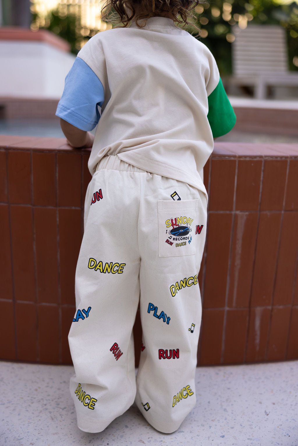 A child wearing a multicolored shirt and cream Sunday Records Pants from the SUNDAY SIBLINGS brand, featuring playful words like "RUN," "DANCE," and "PLAY," stands on a circular brick structure. The twill cotton pants boast colorful embroidery, patch pockets, an elastic waistband, and a patch labeled "SUNDAY" with "Records by Bonnie.