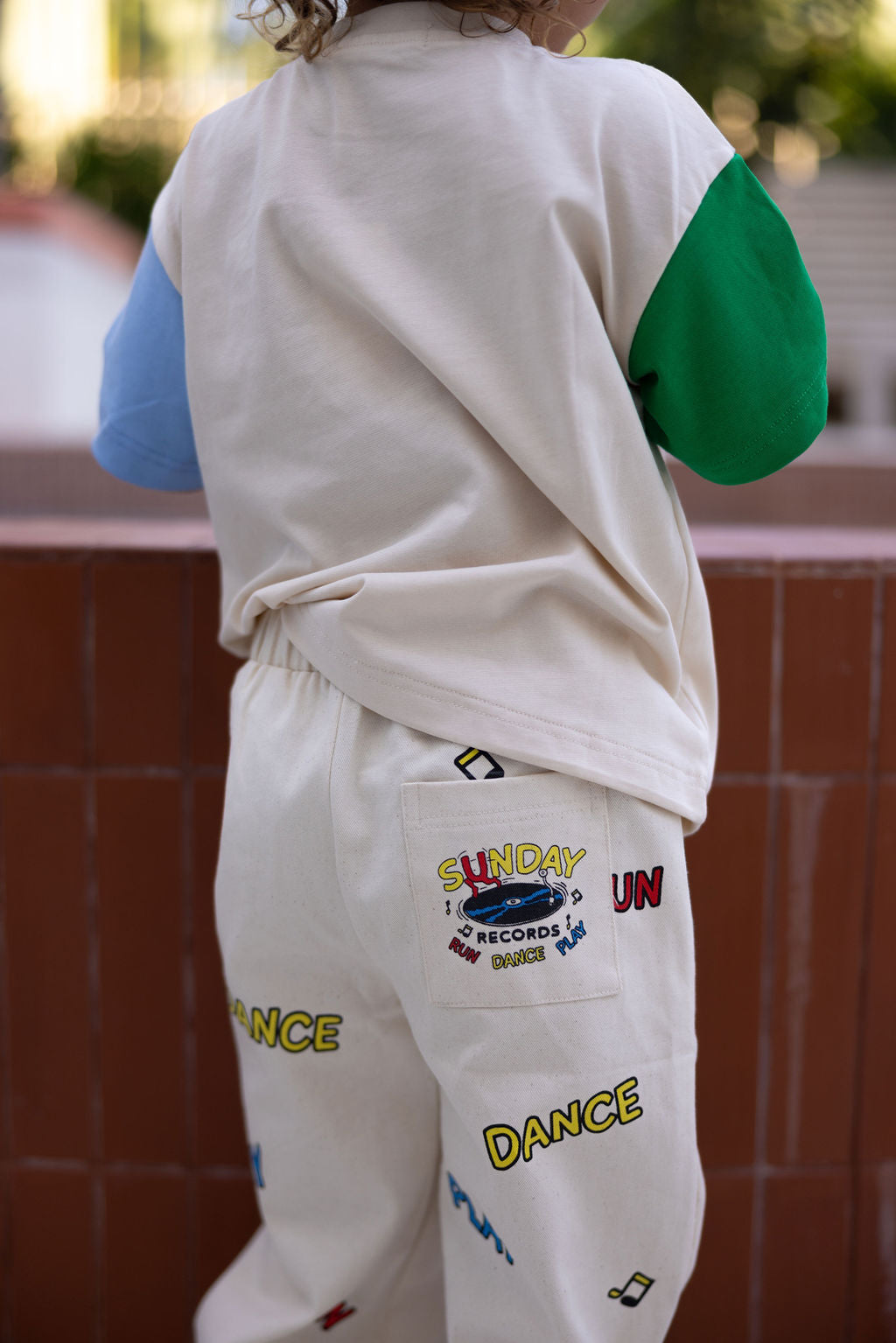 A child is wearing an oversized fit with a blue and green shirt, branded as the Sunday Records Tee by SUNDAY SIBLINGS, paired with cream pants. The pants feature colorful text and graphics such as "Sunday," "Dance," "Records," along with small illustrations on the back pocket, capturing the playful essence reminiscent of Marcus Dixon's designs, near a brick wall outdoors.