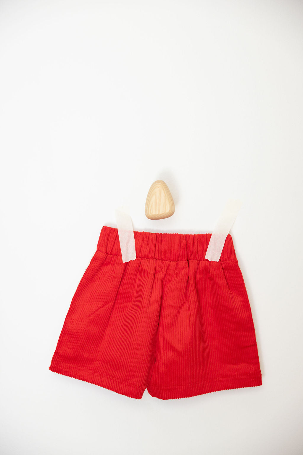 The Playtime Cord Shorts Red by SUNDAY SIBLINGS, featuring an elastic waistband, are displayed on a white wall with a wooden hook, secured by white tape.