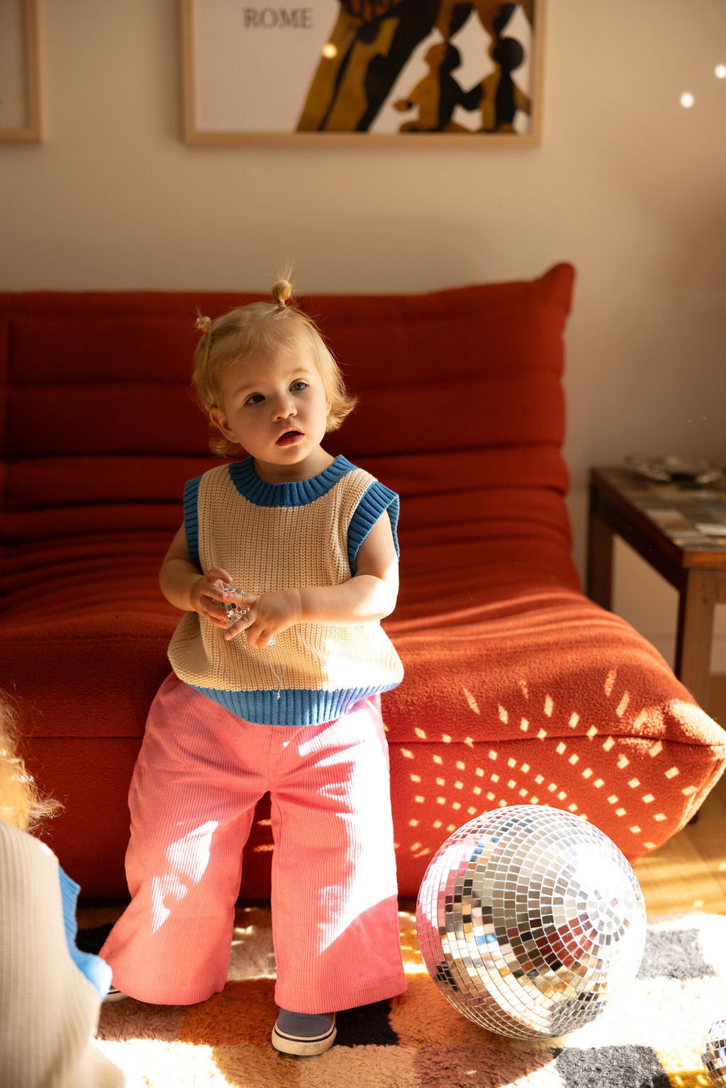 A toddler with short blonde hair is in a sunlit room, holding small objects. They are dressed in a beige Nonno Vest Blue from SUNDAY SIBLINGS featuring blue trim, paired with pink pants, giving off grandad vibes. A disco ball sits on the floor, and a red sofa adds warmth to the scene. The outfit is crafted from 100% cotton for comfort.