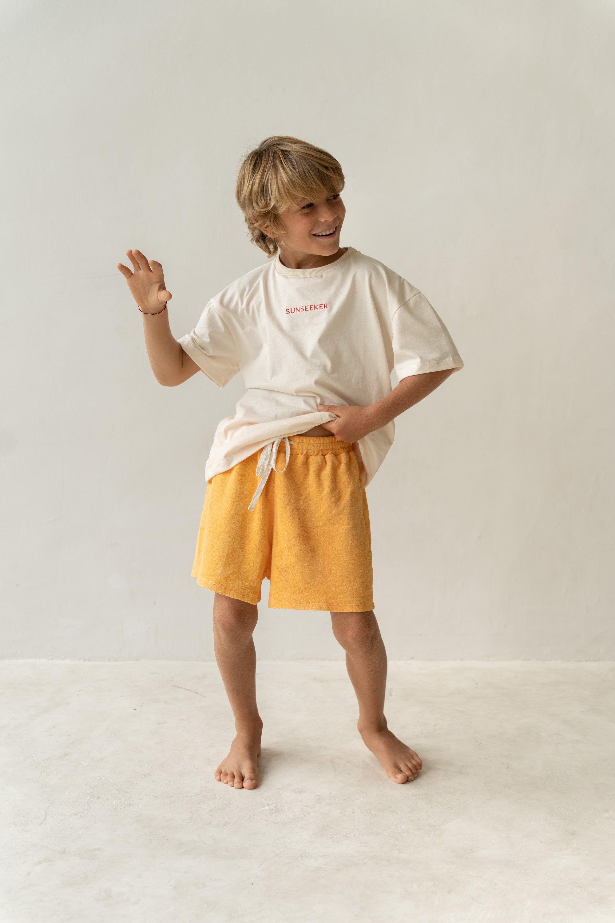 A smiling child with blonde hair stands barefoot, wearing a white T-shirt with red text and Thea Shorts Marigold by ILLOURA THE LABEL, featuring an elasticated waist. One hand is on their hip while the other is raised playfully against a plain light background.