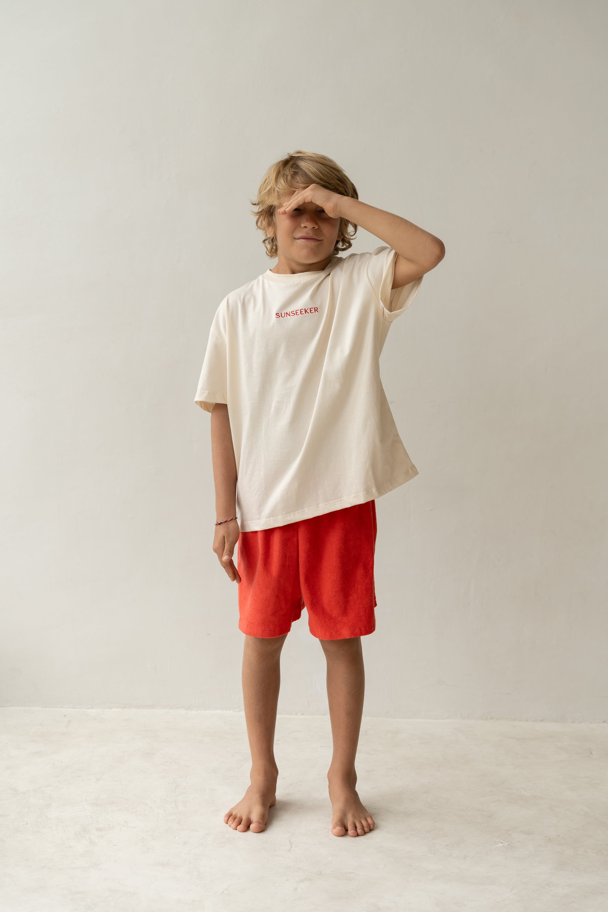 A barefoot child with blonde hair stands on a light floor in a cream "SUNSEEKER" T-shirt and vibrant red Watermelon Thea Shorts by ILLOURA THE LABEL, featuring a cotton terry towelling fabric and elasticated waist. They shield their eyes with their right hand, posing against a plain white wall.