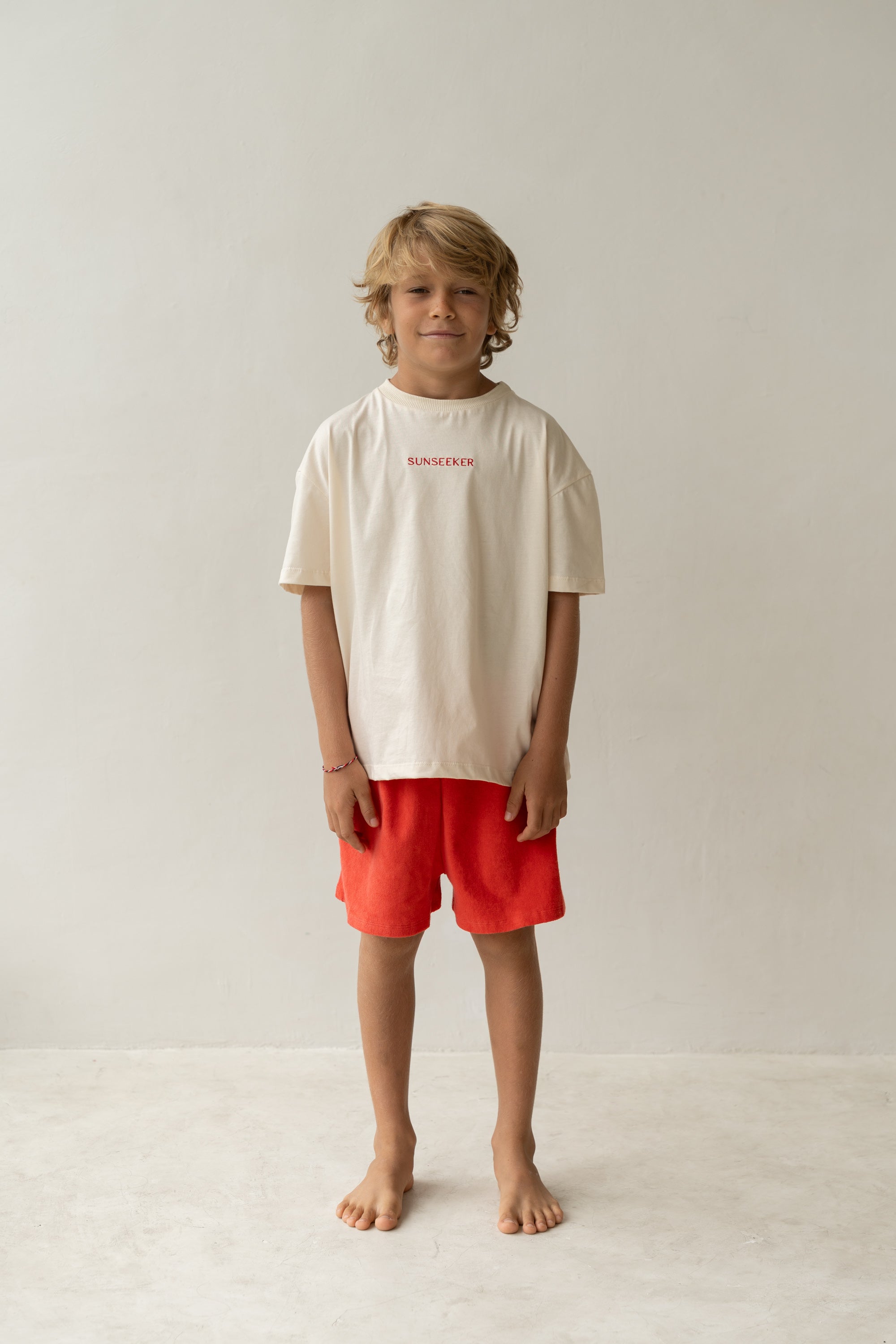 A smiling child with tousled blonde hair stands barefoot against a plain background, wearing a white T-shirt and bright red Thea Shorts Watermelon from ILLOURA THE LABEL. The shorts feature an elasticated waist for comfort, and the shirt proudly displays the word "SUNSCREEN.