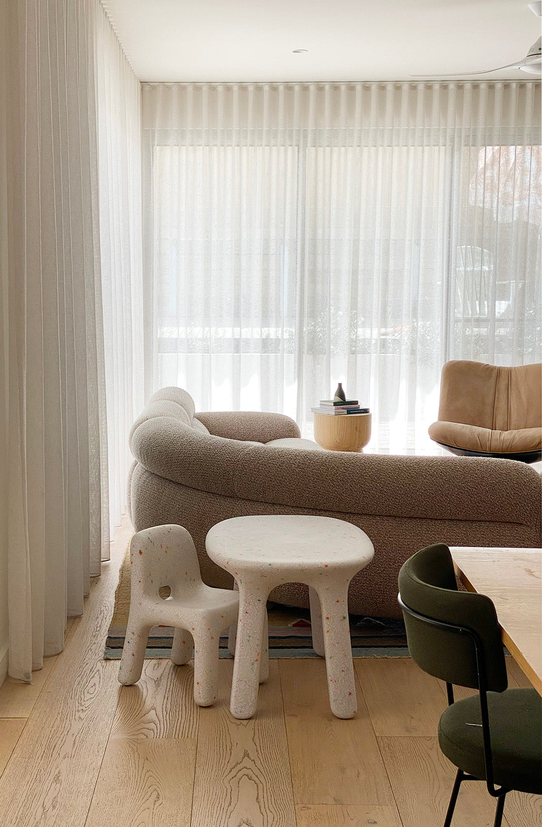 A stylish living room with large windows covered by sheer white curtains, allowing soft light to filter through. It features a modern beige curved sofa, a small BAY KIDS Kid's Table and Chair Set - perfect for kids' activities - a tan armchair, and light wooden flooring with a subtle rug.