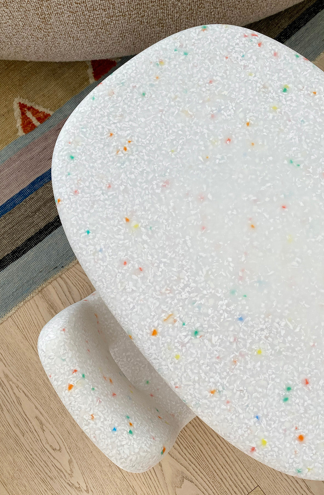 A rounded white table from the BAY KIDS Kid's Table and Chair Set is pictured. The table boasts a speckled, terrazzo-like surface adorned with colorful spots of orange, green, blue, and yellow. Made from recycled plastic furniture, it is situated on a wooden floor with part of a beige couch and a rug visible in the background.