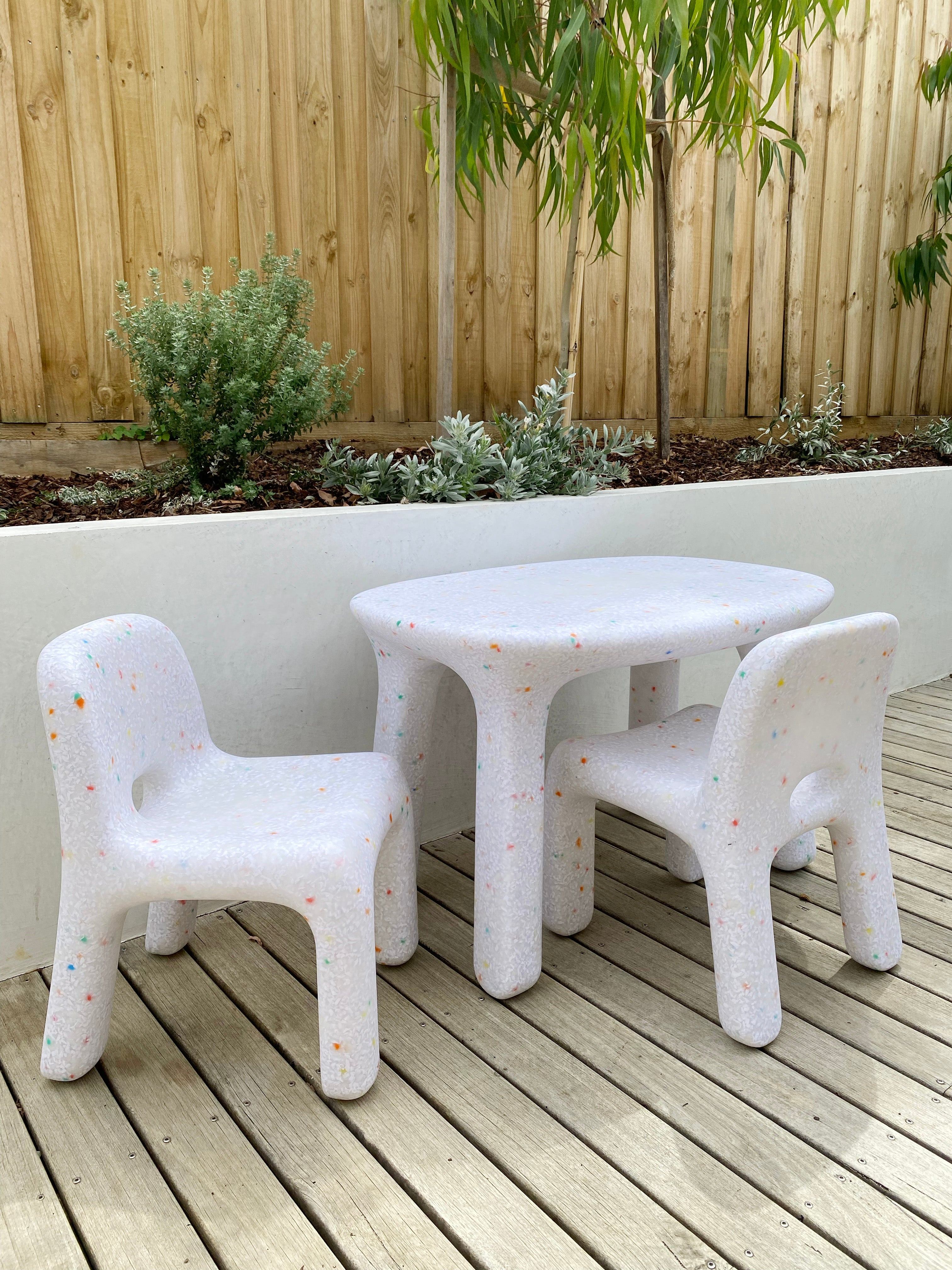 A charming, small BAY KIDS table and chair set in white, featuring colorful speckles, adorns the wooden deck. Designed specifically for outdoor use, this Kid's Table and Chair Set is positioned against a white wall with leafy green plants and a wooden fence as the backdrop.