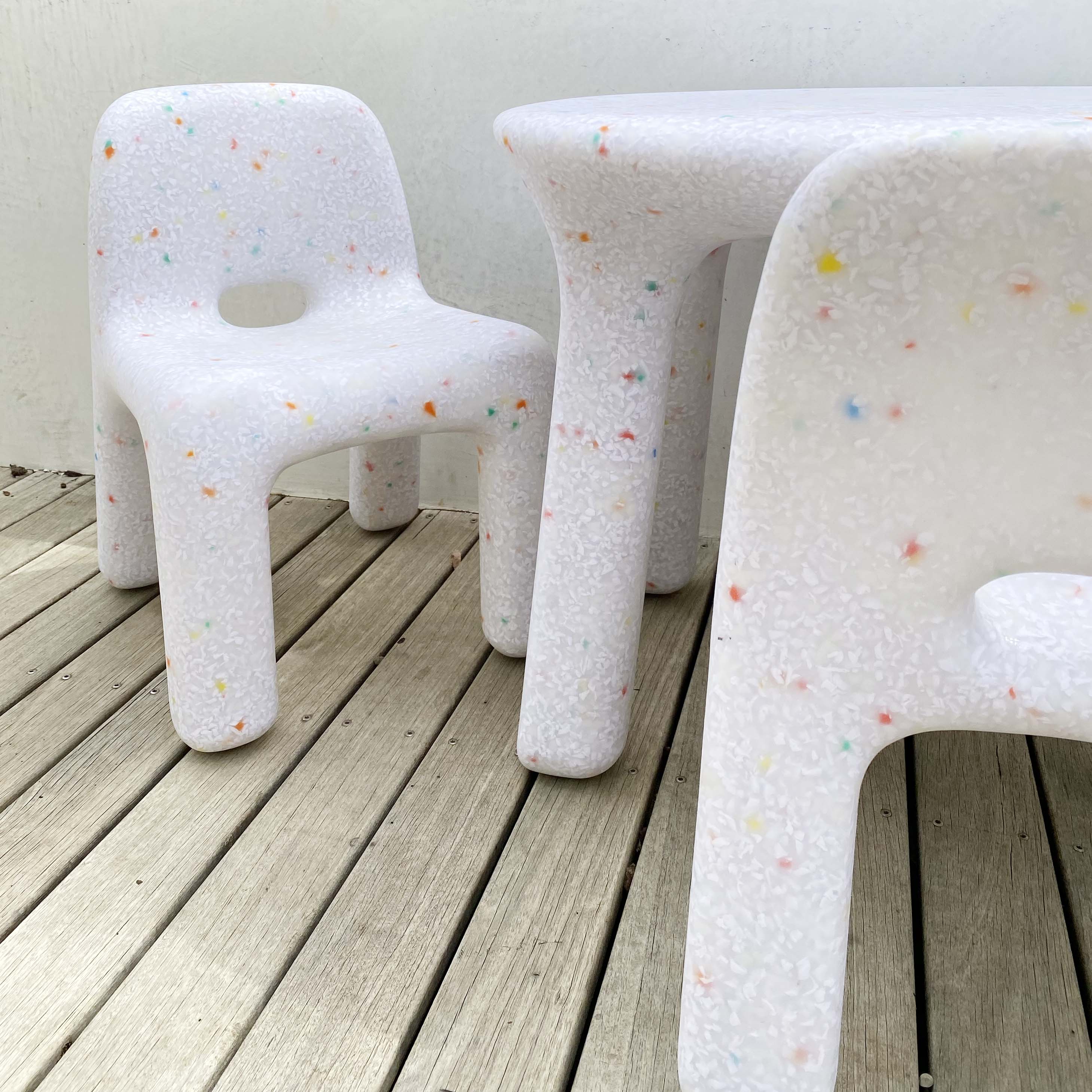 A close-up view of the BAY KIDS Kid's Table and Chair Set, crafted from speckled white recycled plastic material. The chunky, rounded design makes it ideal for outdoor use, while the colorful speckles add a playful touch. The furniture is charmingly placed on a wooden deck.