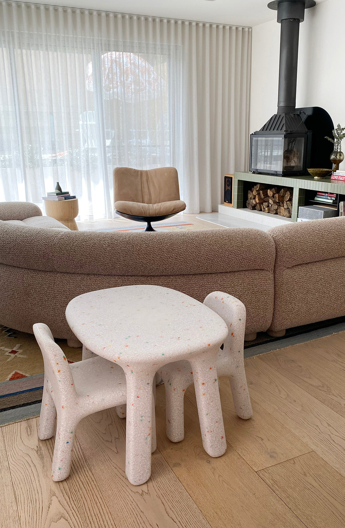 A modern living room with light wood flooring and neutral tones features a plush, curved beige sofa, a tan swivel chair, and a wood-burning stove. In the foreground, the BAY KIDS Kid's Table and Chair Set made from speckled white recycled plastic adds an eco-friendly touch to the decor.