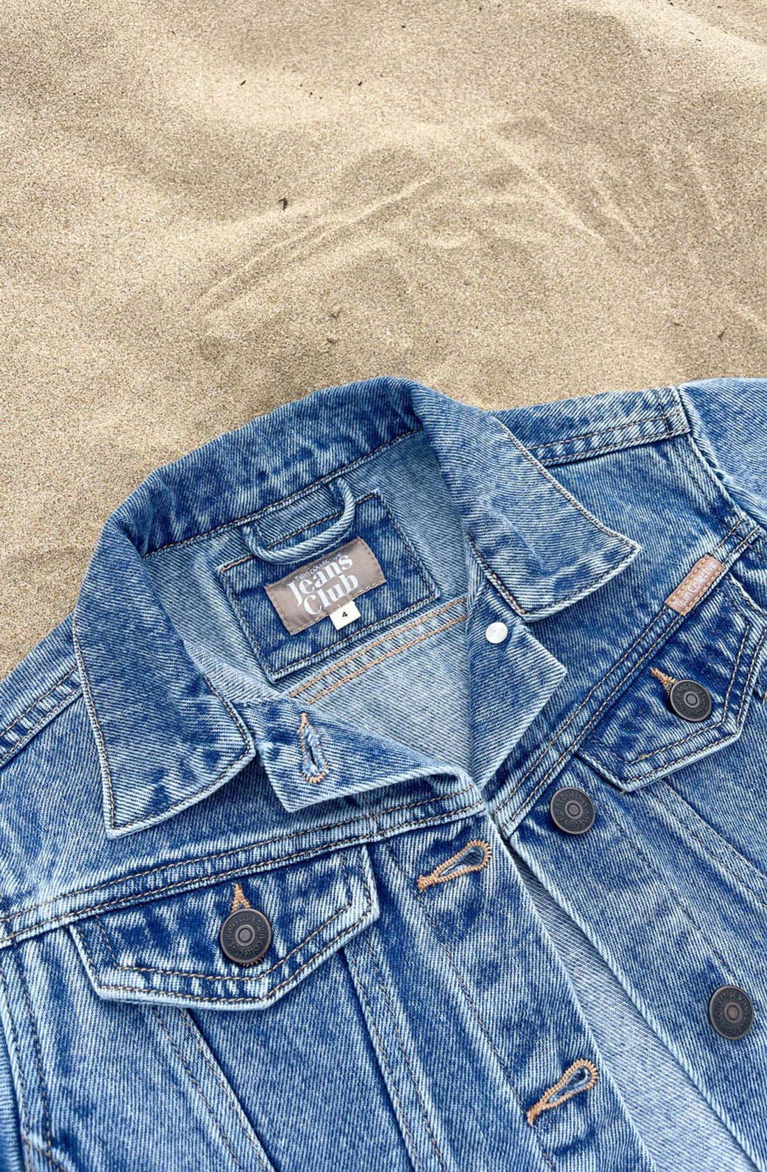 A Trucker Jacket Thunder Blue, featuring metal buttons and evoking a 1990s-inspired vintage denim style, is laid out on the sand. The label on the inside collar reads "TWIN COLLECTIVE" with the size "Large.