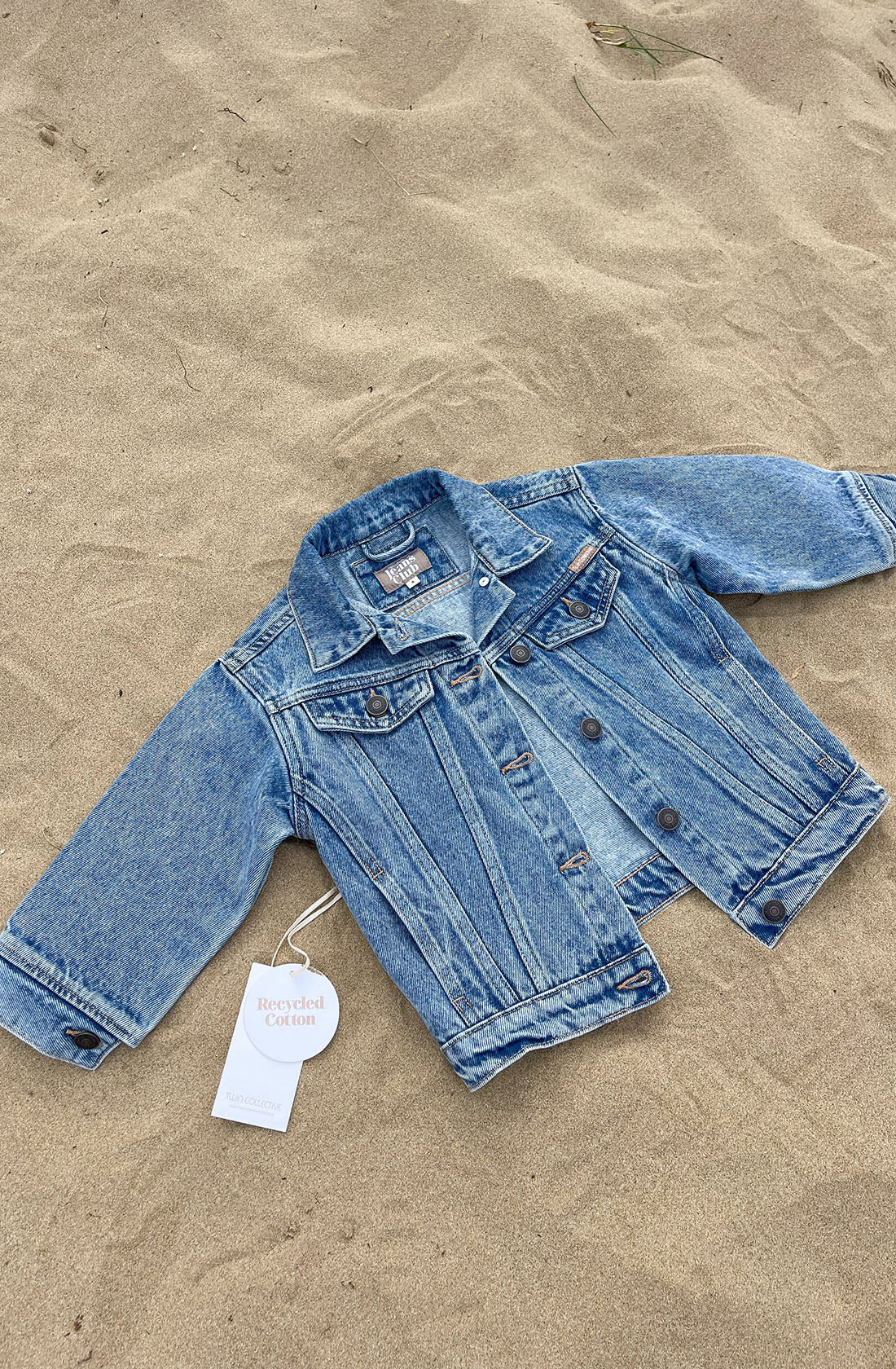 The Trucker Jacket Thunder Blue by TWIN COLLECTIVE is displayed on a sandy surface. This light blue denim jacket features long sleeves, metal buttons, and two chest pockets. An attached tag reads "Recycled Cotton," highlighting its sustainable cotton origins. The piece exudes a 1990s-inspired, vintage denim jackets vibe.