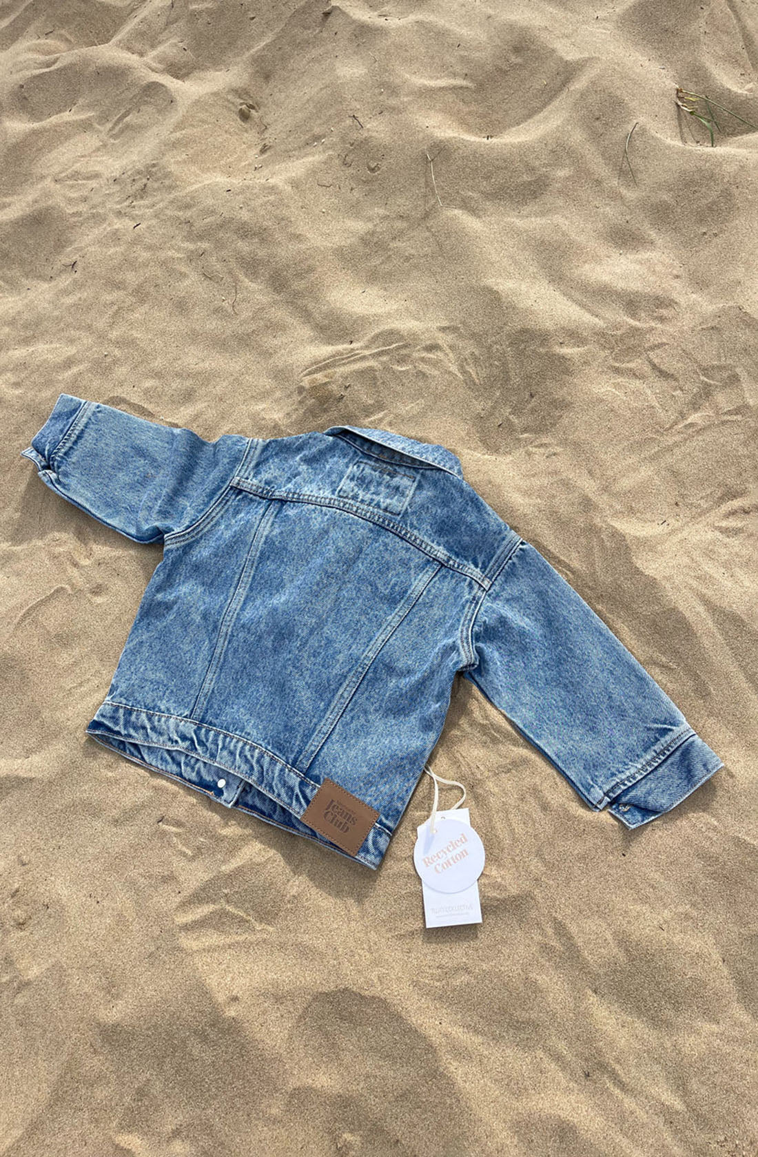 A small, 1990s-inspired Trucker Jacket in Thunder Blue from TWIN COLLECTIVE is spread out on sandy ground. A white price tag attached to the jacket displays text, and a small brown tag is also visible. The jacket appears to be new, with labels still attached.