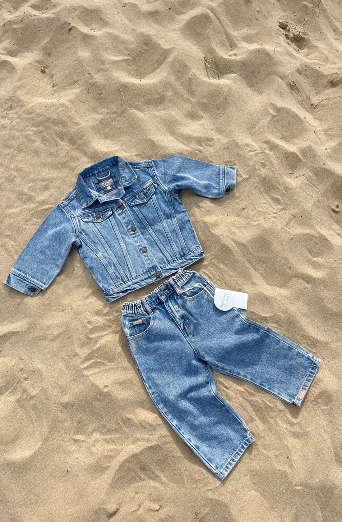 A children's Trucker Jacket Thunder Blue and matching elastic-waist pants by TWIN COLLECTIVE are laid out on sandy ground. The outfit includes a button-up, long-sleeved jacket and pants, both crafted from sustainable cotton in a blue washed vintage denim texture reminiscent of 1990s-inspired styles.