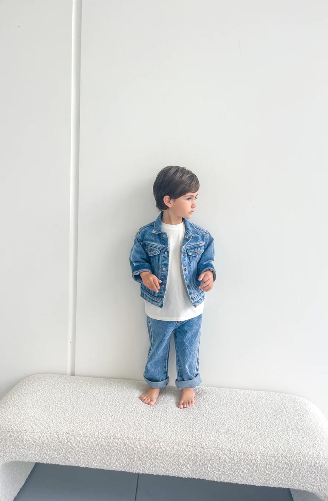 A young child stands barefoot on a white textured bench against a plain white wall, wearing the TWIN COLLECTIVE Trucker Jacket Thunder Blue layered over a white shirt and sustainable denim pants, looking to the side.