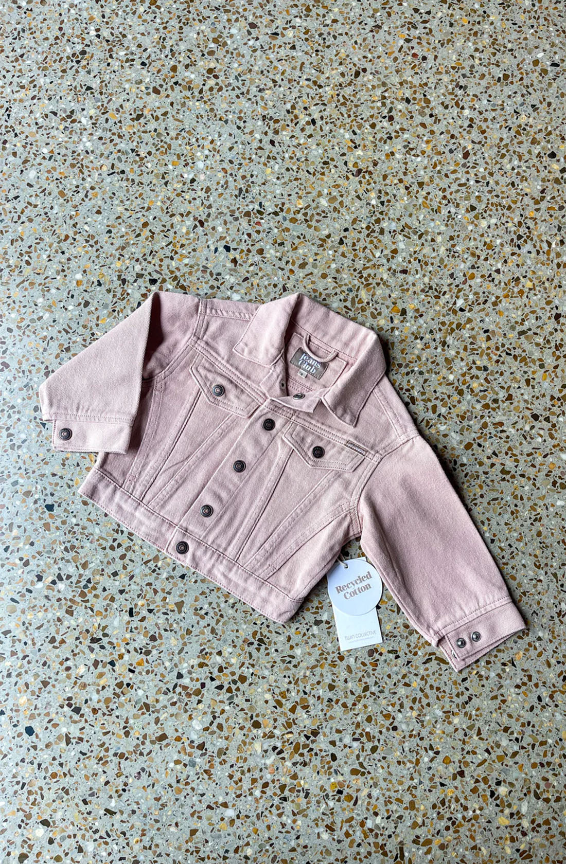 The Trucker Jacket Dusty Pink by TWIN COLLECTIVE, designed for toddlers and young children, is laid flat on a speckled terrazzo floor. Featuring metal buttons and chest pockets, this 1990s-inspired fashion piece is crafted from sustainable cotton denim with a tag attached, indicating it is brand new.