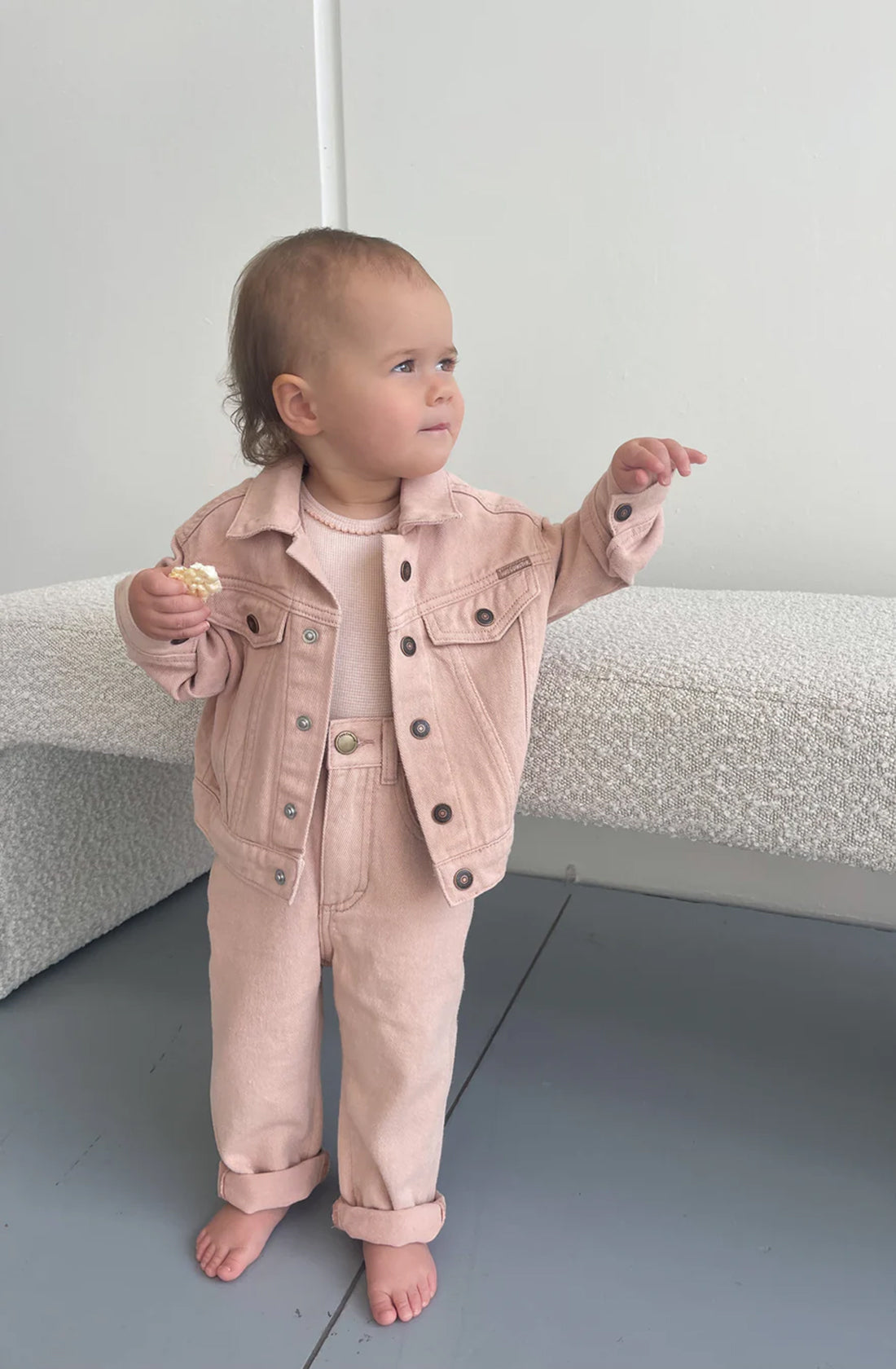 A toddler stands barefoot on the floor in a minimalistic room, wearing a light pink Trucker Jacket Dusty Pink from TWIN COLLECTIVE and matching pants made of sustainable cotton denim. The child holds a small snack in one hand and appears to be pointing or reaching out with the other hand, looking off to the side.