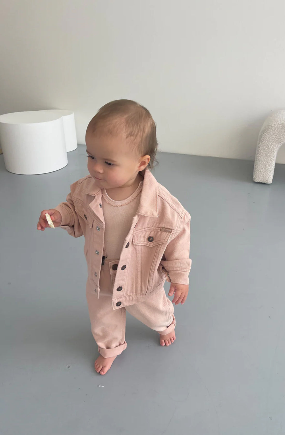 A toddler with light brown hair wearing a TWIN COLLECTIVE Trucker Jacket in Dusty Pink and matching pants stands indoors on a grey floor. The child is barefoot, holding a small object in their right hand. Minimalist white furniture is in the background, giving a nod to 1990s-inspired fashion with its simple elegance.