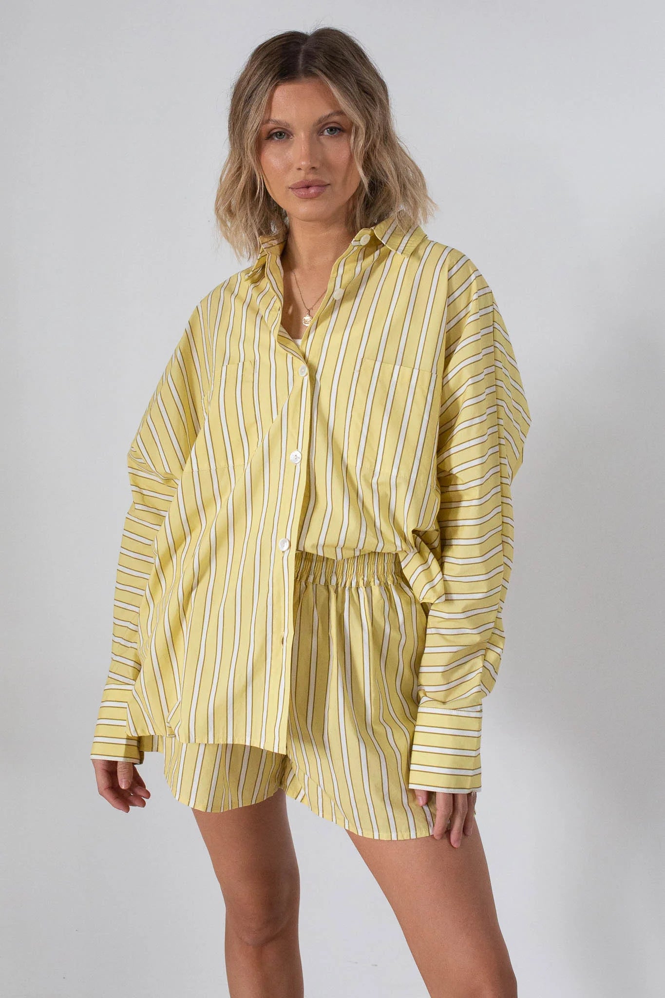 A woman wears the Tiny Trove - Women's Isla Stripe Cotton Set Yellow/White Stripe by TINY TROVE, featuring a relaxed-fit, striped button-up shirt and matching elastic-waist shorts, standing against a plain light background.