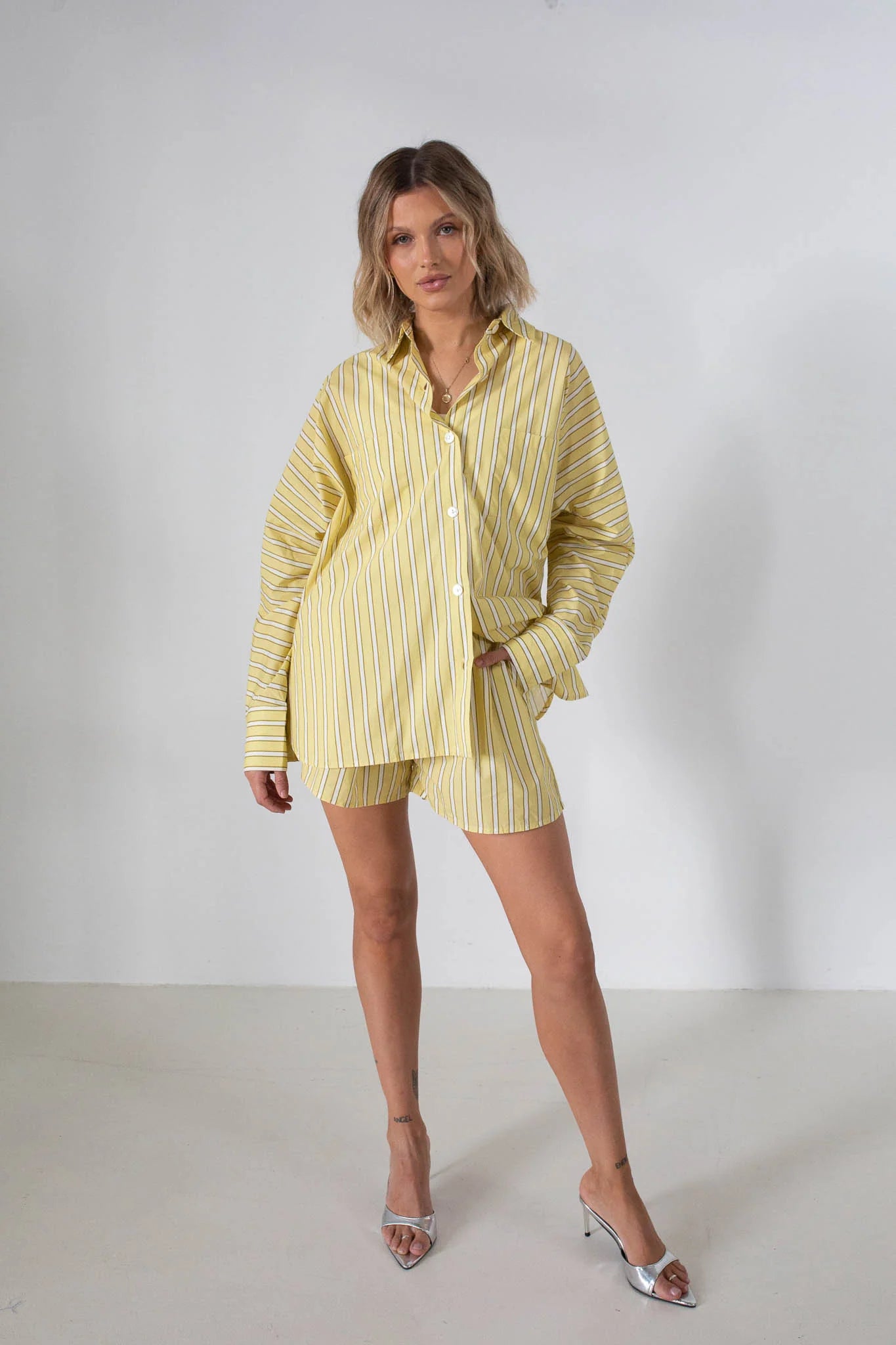 A woman stands against a white background wearing TINY TROVE’s Isla Stripe Cotton Set in yellow and white stripes, featuring a relaxed-fit button-up shirt and matching shorts, styled with silver high-heeled sandals.