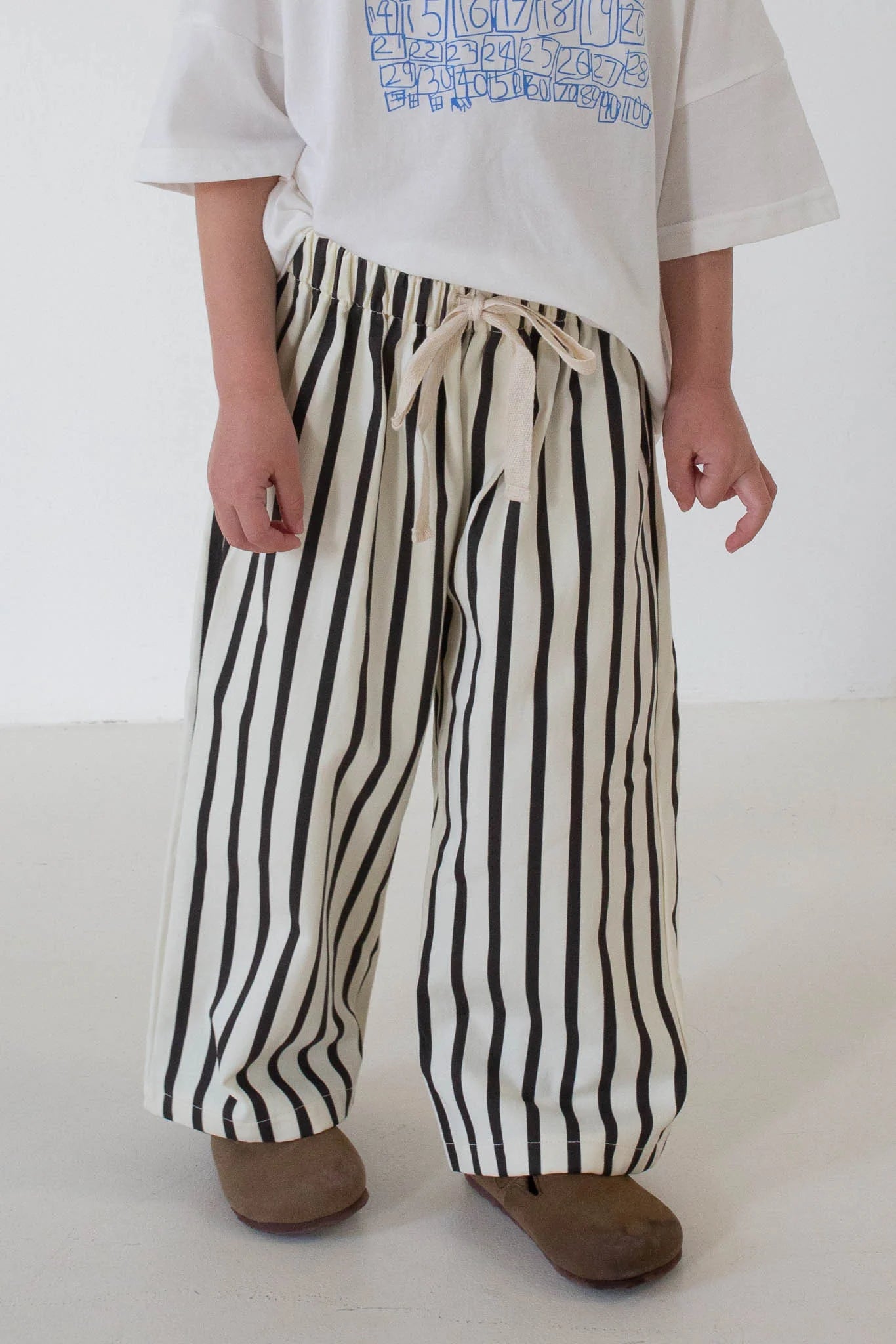 A child in Tiny Trove - Billie Stripe Cotton Pants Cream/Cocoa Stripe by TINY TROVE, styled with a white graphic tee and brown shoes, stands against a plain light background.