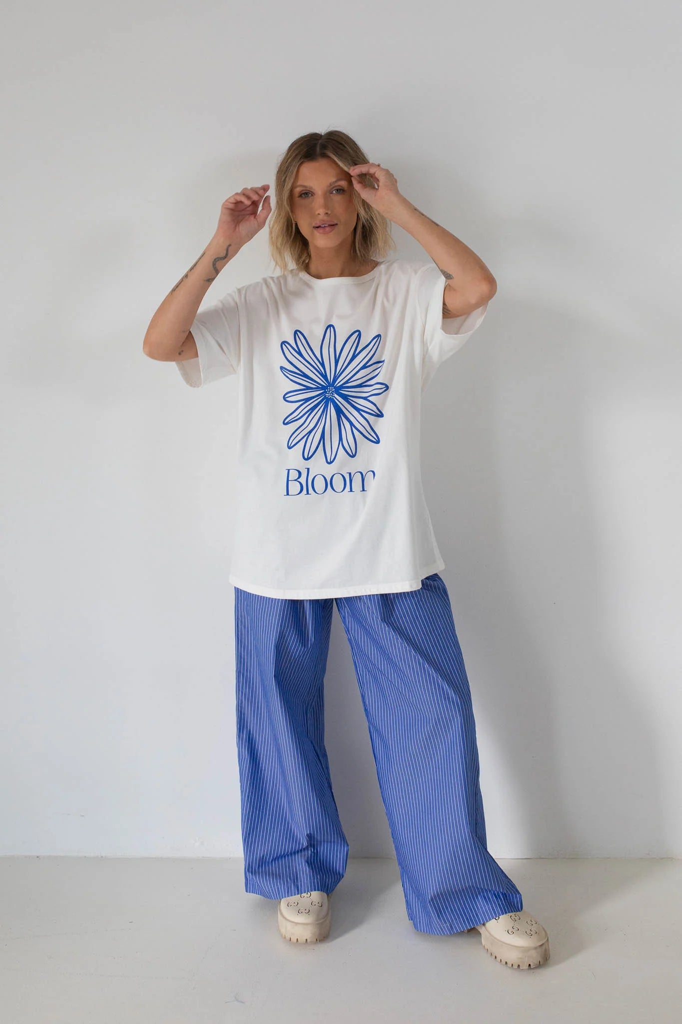 A person stands against a plain white wall wearing the TINY TROVE Tiny Trove - Women's Bloom Relaxed Tee White/Blue, featuring a blue flower graphic and “Bloom,” paired with blue striped wide-leg pants and light-colored platform shoes.