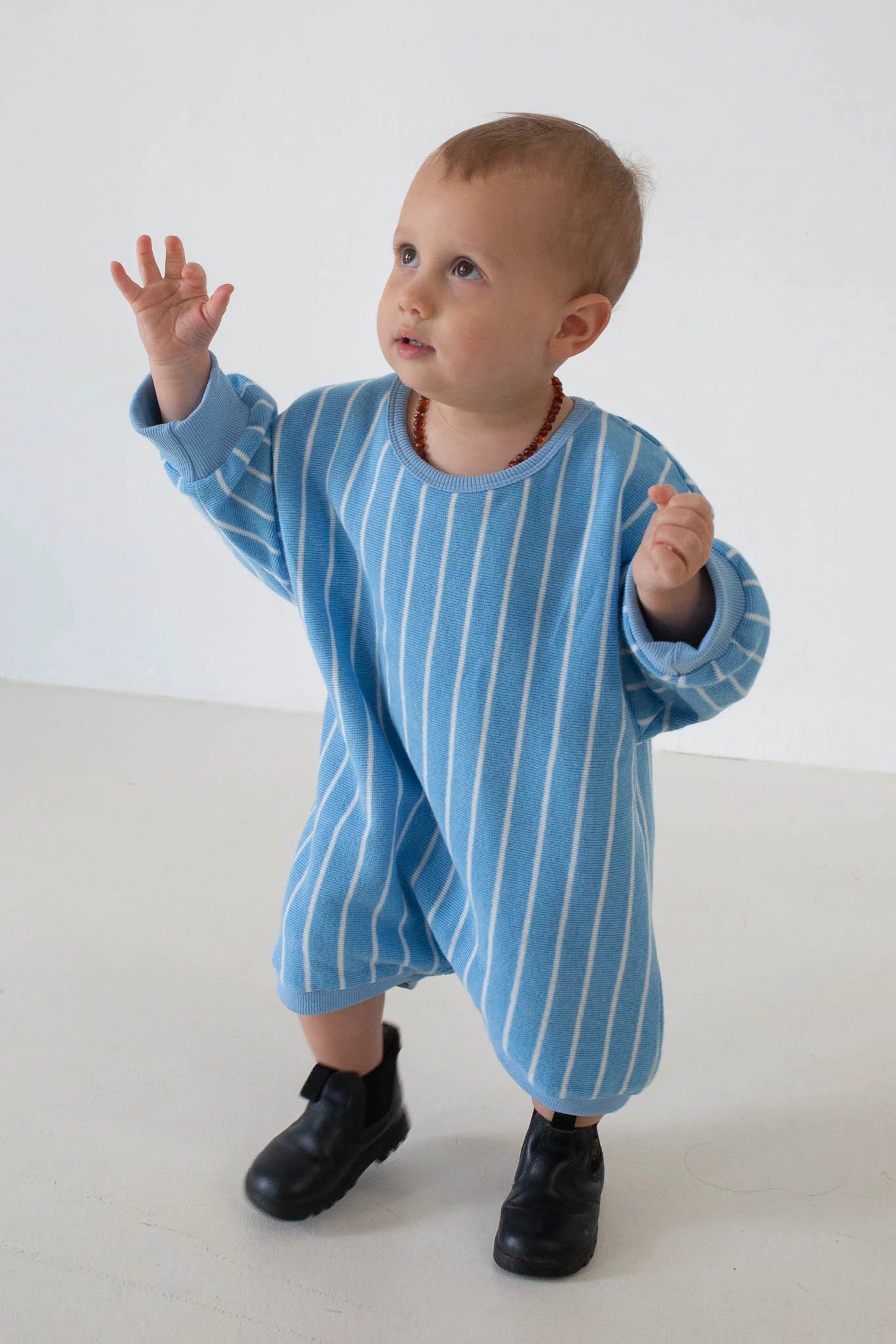 A toddler wears the TINY TROVE Addie Cotton Stripe Romper in blue and white stripes, paired with black boots and a beaded necklace, standing on a light floor and looking up with one hand raised.