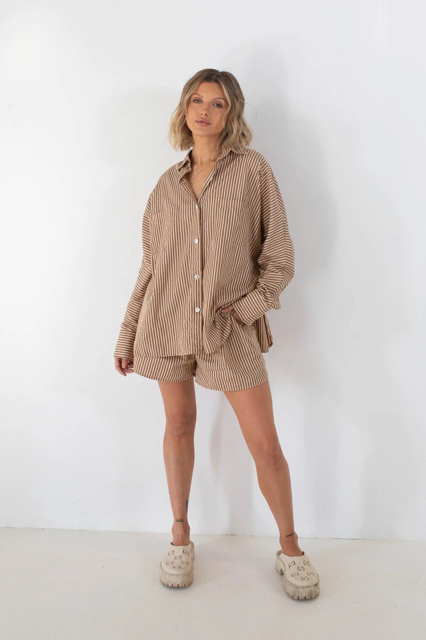 A woman stands against a white wall wearing the TINY TROVE Isla Stripe Cotton Set in Caramel/White Stripe, an oversized shirt and matching shorts, paired with beige slip-on shoes.