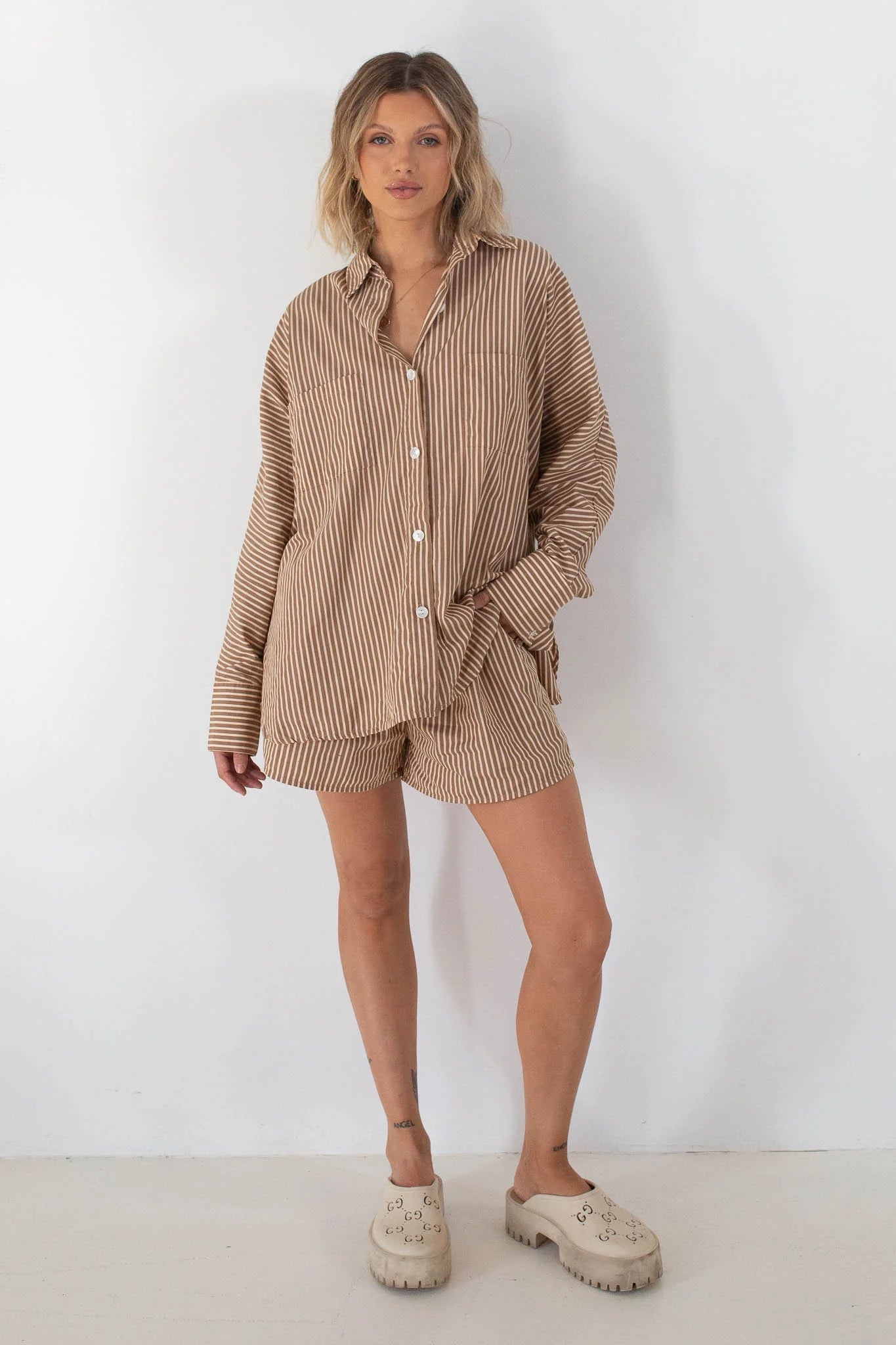 A woman with blonde hair stands against a white wall wearing the TINY TROVE - Women's Isla Stripe Cotton Set in Caramel/White Stripe, featuring an oversized button-up shirt and matching shorts, paired with light chunky clogs.
