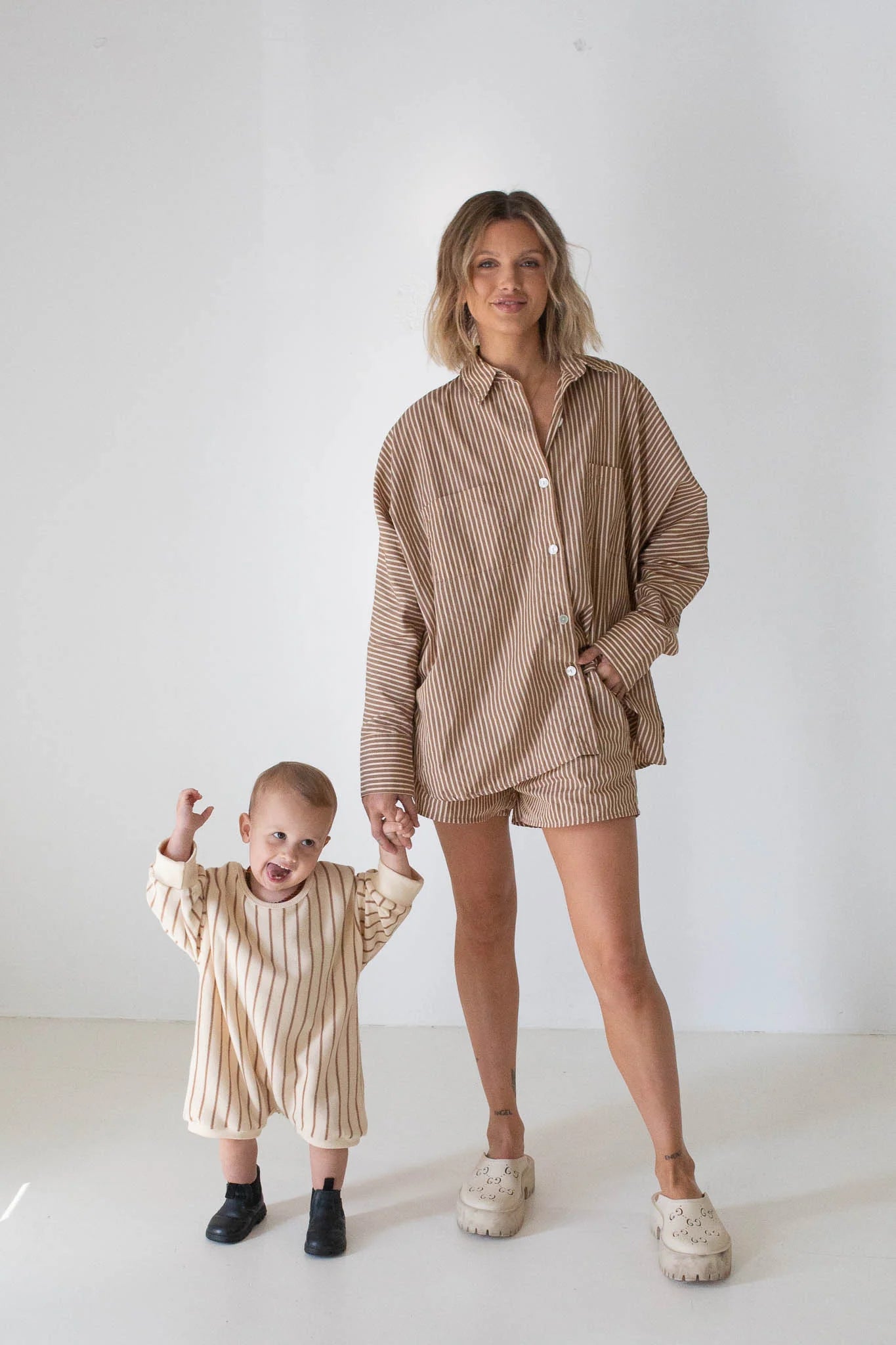 A woman wears the TINY TROVE Isla Stripe Cotton Set in Caramel/White Stripe, while a small child in a cream and brown striped outfit with black shoes holds her hand. Both stand against a white background, smiling.