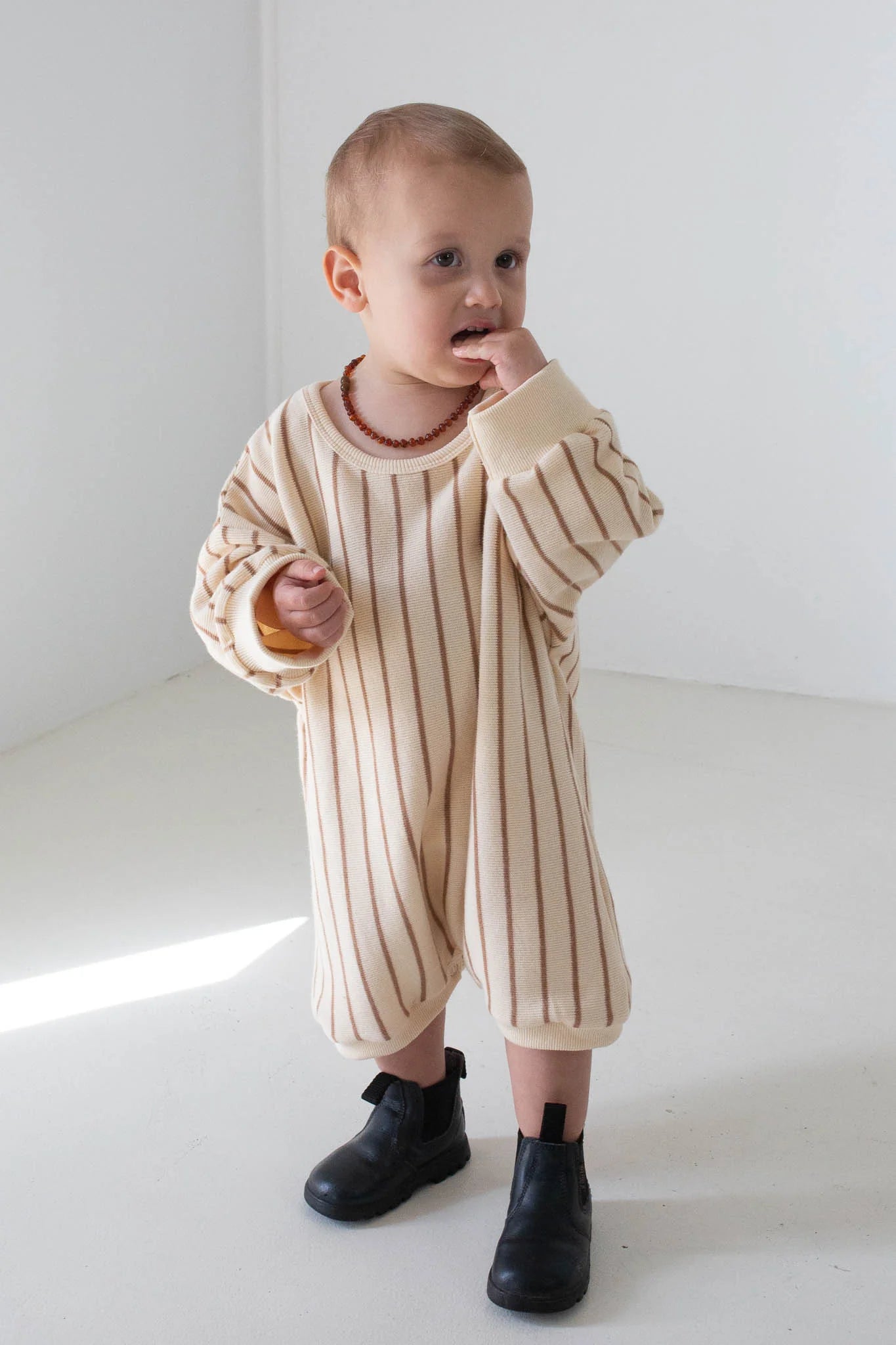 A toddler stands in a minimal white room, wearing the TINY TROVE Addie Cotton Stripe Romper in Cream/Latte Stripe with black boots, one hand in their mouth.