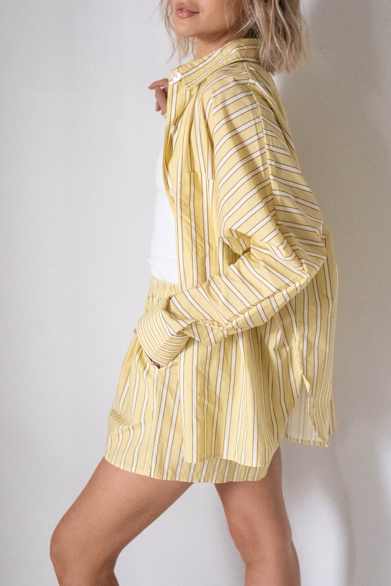 A woman poses sideways in the TINY TROVE - Women's Isla Stripe Cotton Set Yellow/White Stripe, featuring a relaxed-fit striped button-up shirt and matching shorts over a white top, with one hand in her pocket against a plain background.